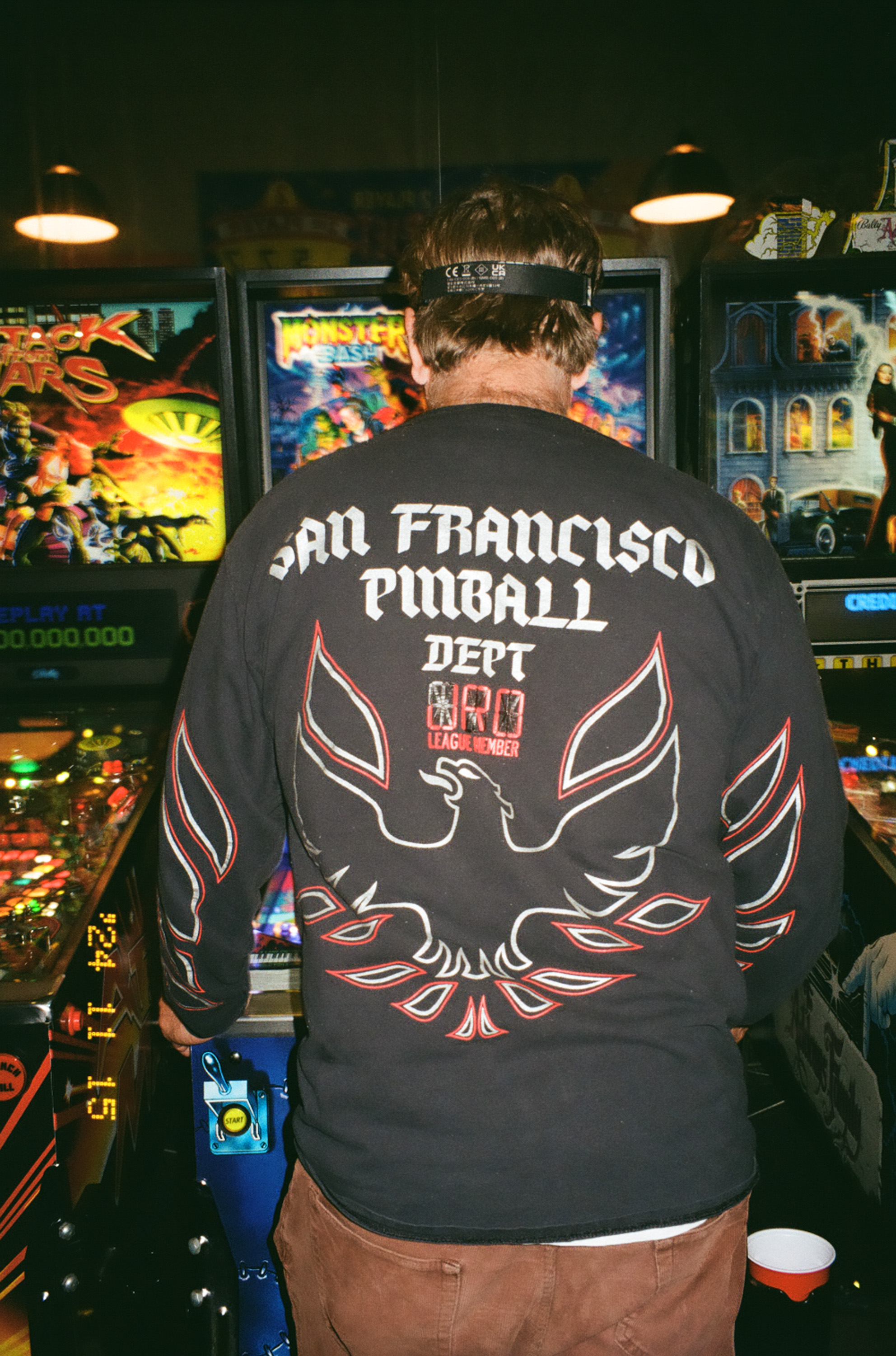 A person in a &quot;San Francisco Pinball Dept&quot; shirt stands in front of pinball machines with brightly lit designs, focused intently on the game.