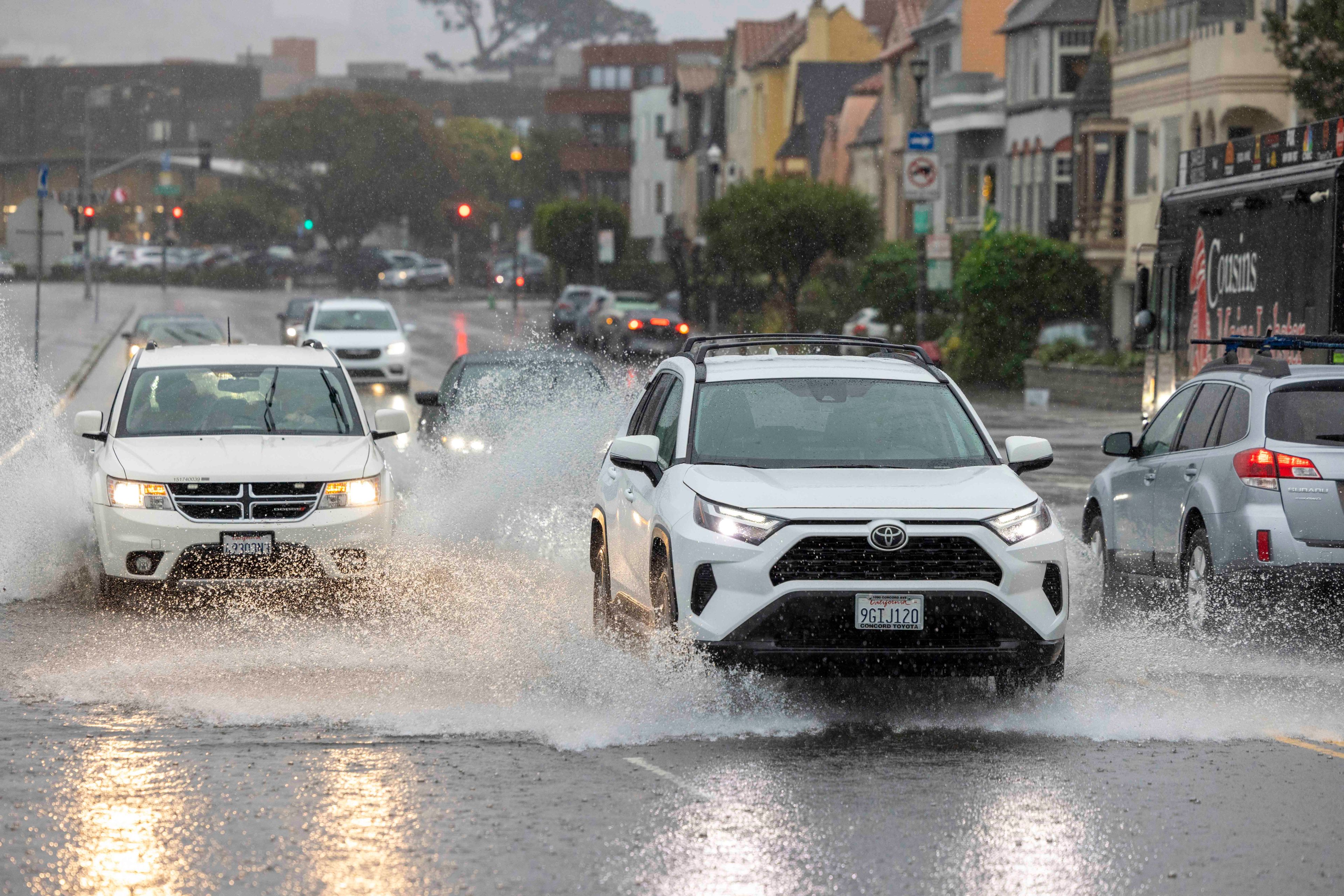 Cars drive through heavy rain and water splashes on a flooded street in a city, with buildings and traffic lights in the background.