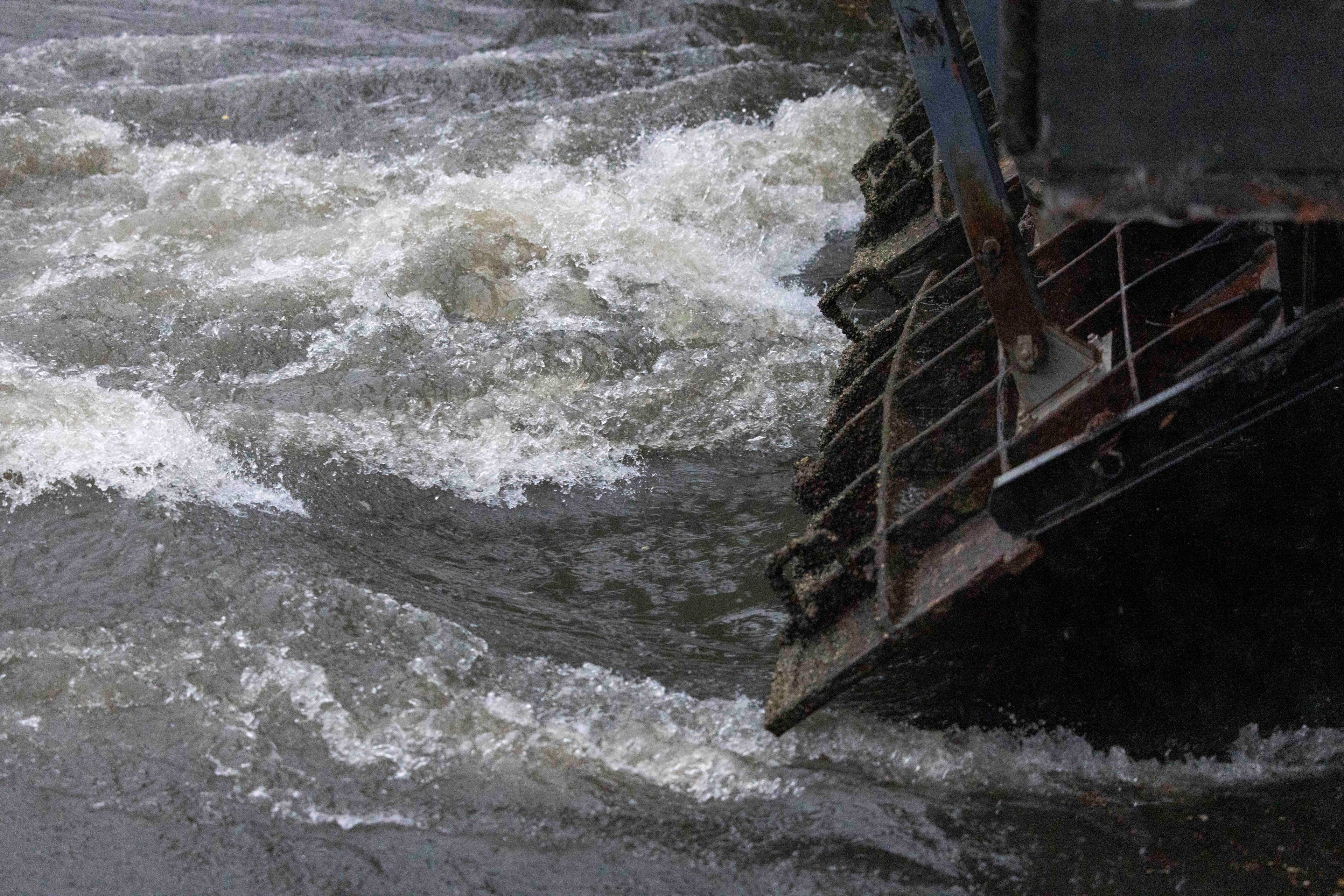 The image shows a turbulent river with foamy white water swirling around a partially submerged metal waterwheel.