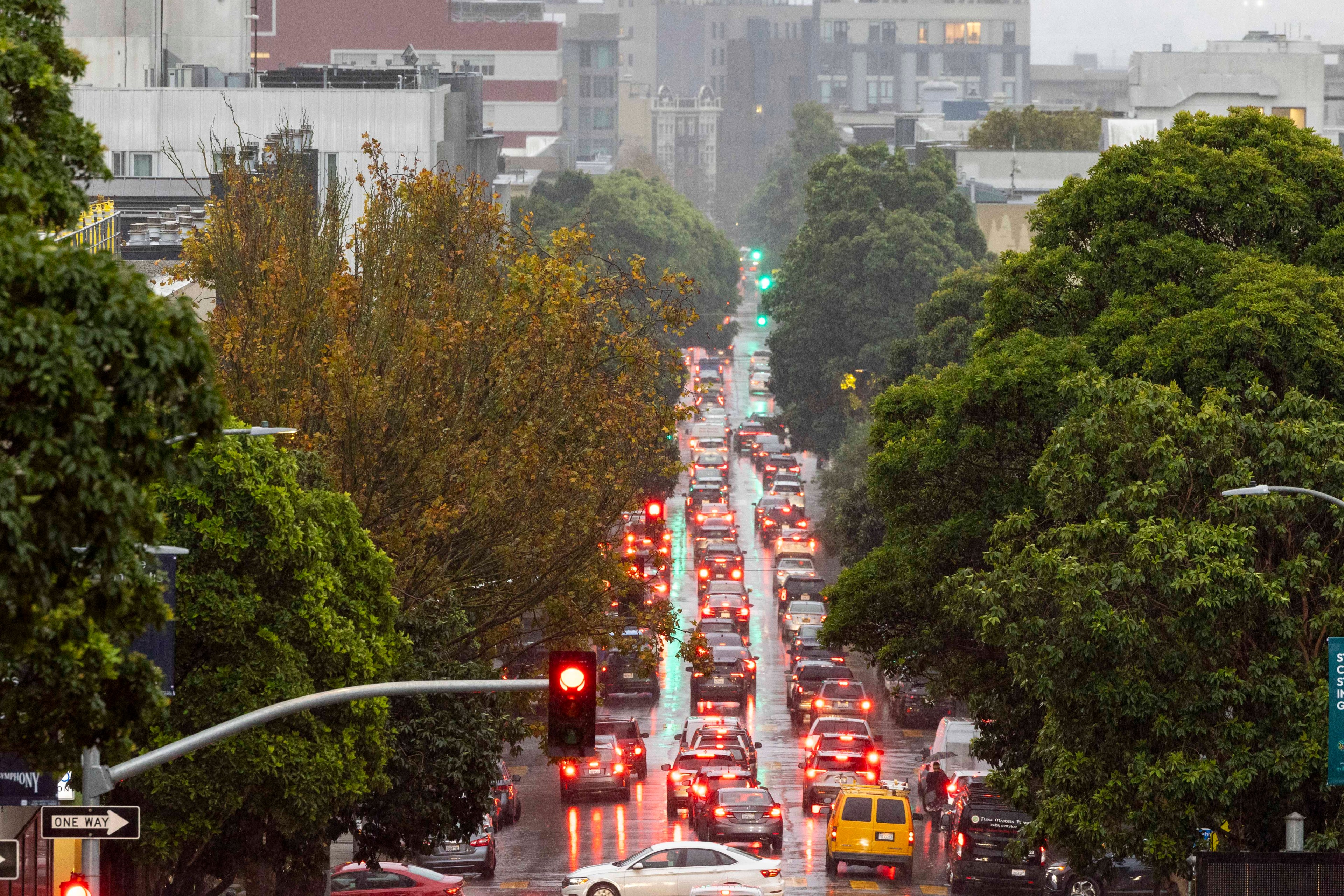 A rainy urban street densely packed with cars and glowing brake lights stretches between lush green trees and buildings under a cloudy sky.