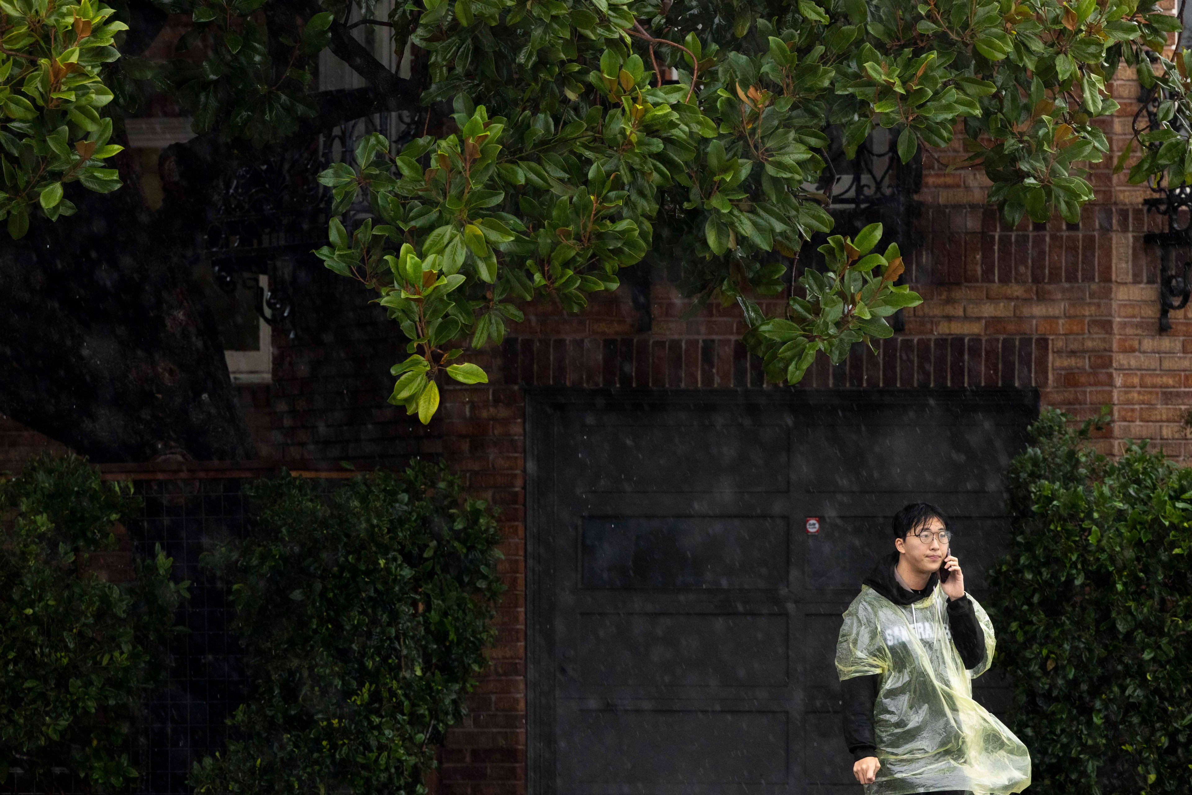 A person in a yellow rain poncho stands under a tree during a rain shower, talking on a phone. They're beside a brick building with a dark garage door.