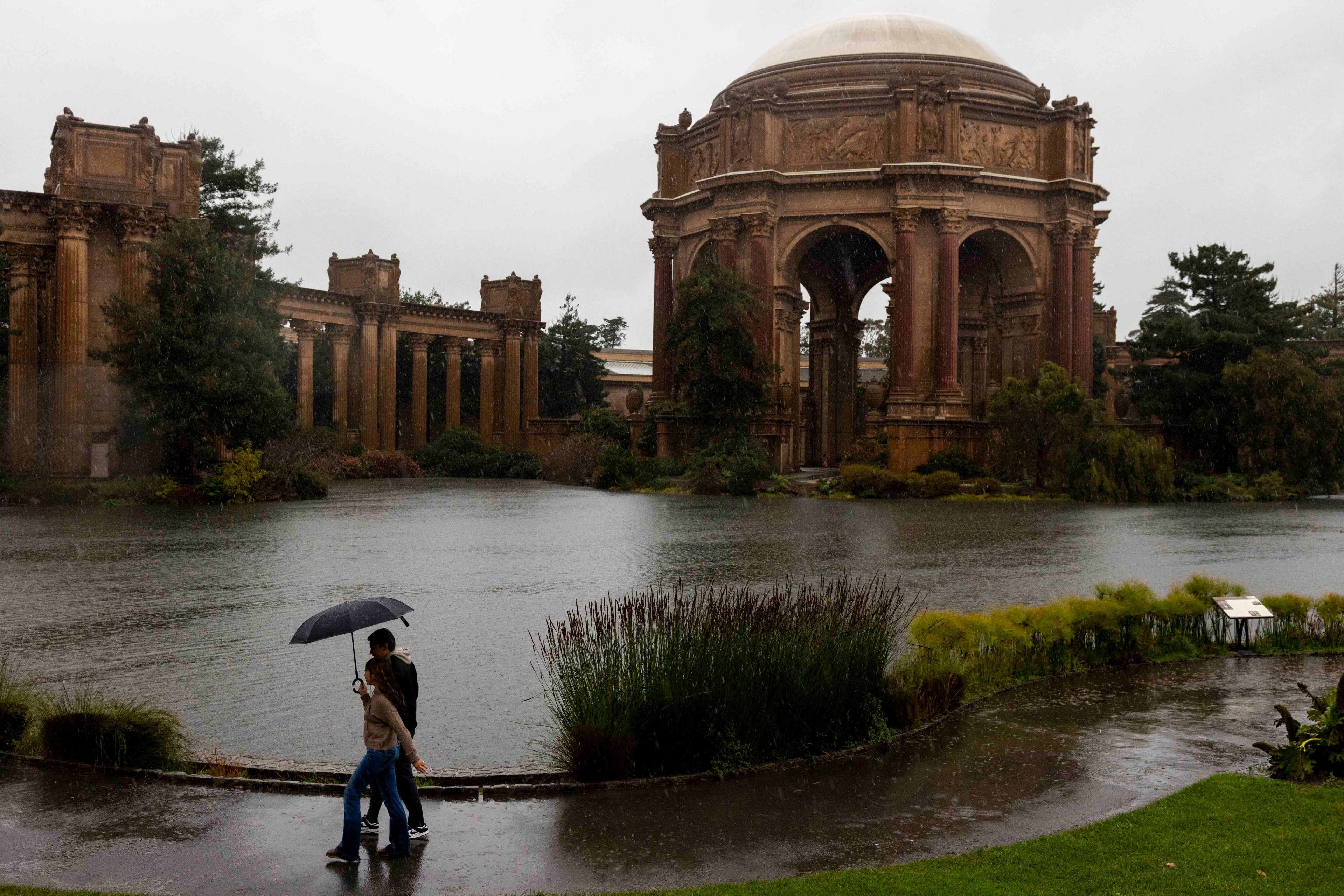 Two people with an umbrella walk along a path by a reflective pond. In the background is a grand, domed classical structure surrounded by greenery.