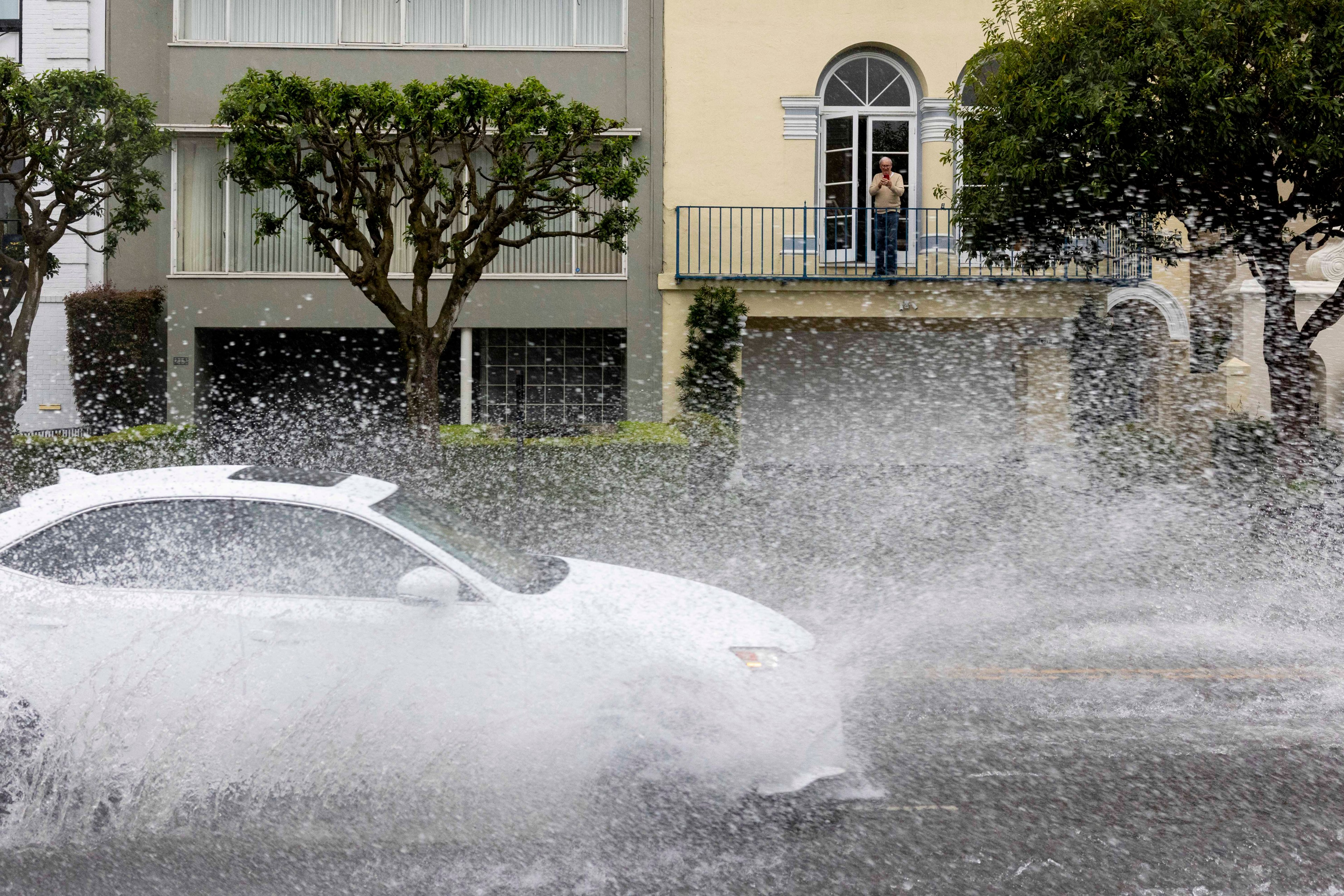 A white car speeds through a large puddle, creating a big splash of water. A person observes the scene from a balcony in front of a building with trees nearby.