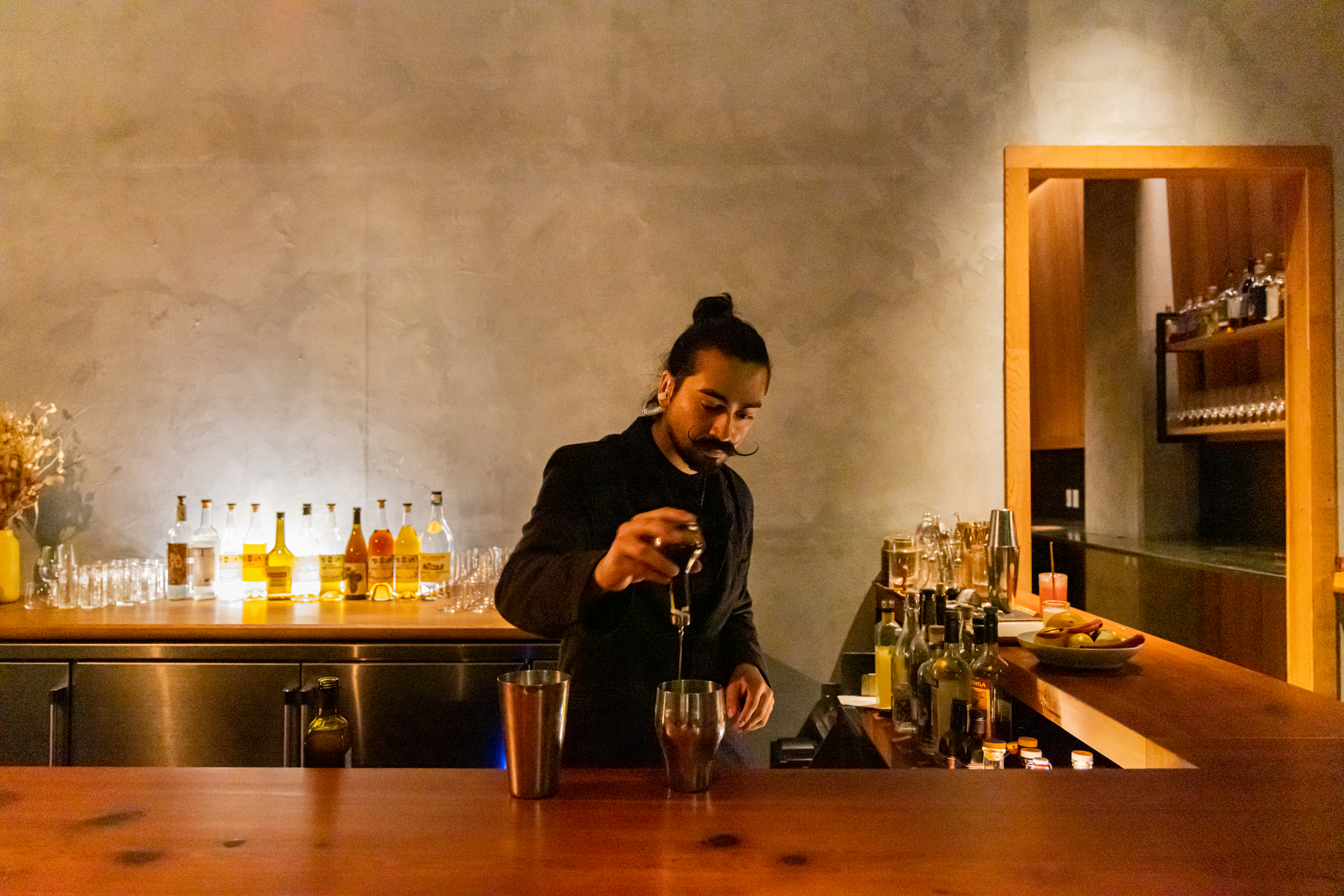 A bartender with a mustache and topknot pours a drink behind a well-lit bar, surrounded by bottles and glasses on a wooden counter in a cozy setting.