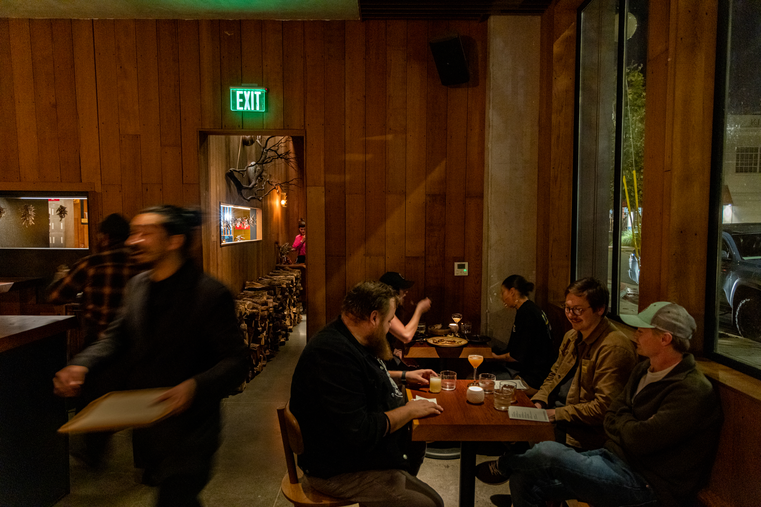 A cozy restaurant with wooden walls, featuring a group of people laughing at a table. A server walks by, and dim lighting adds a warm ambiance.