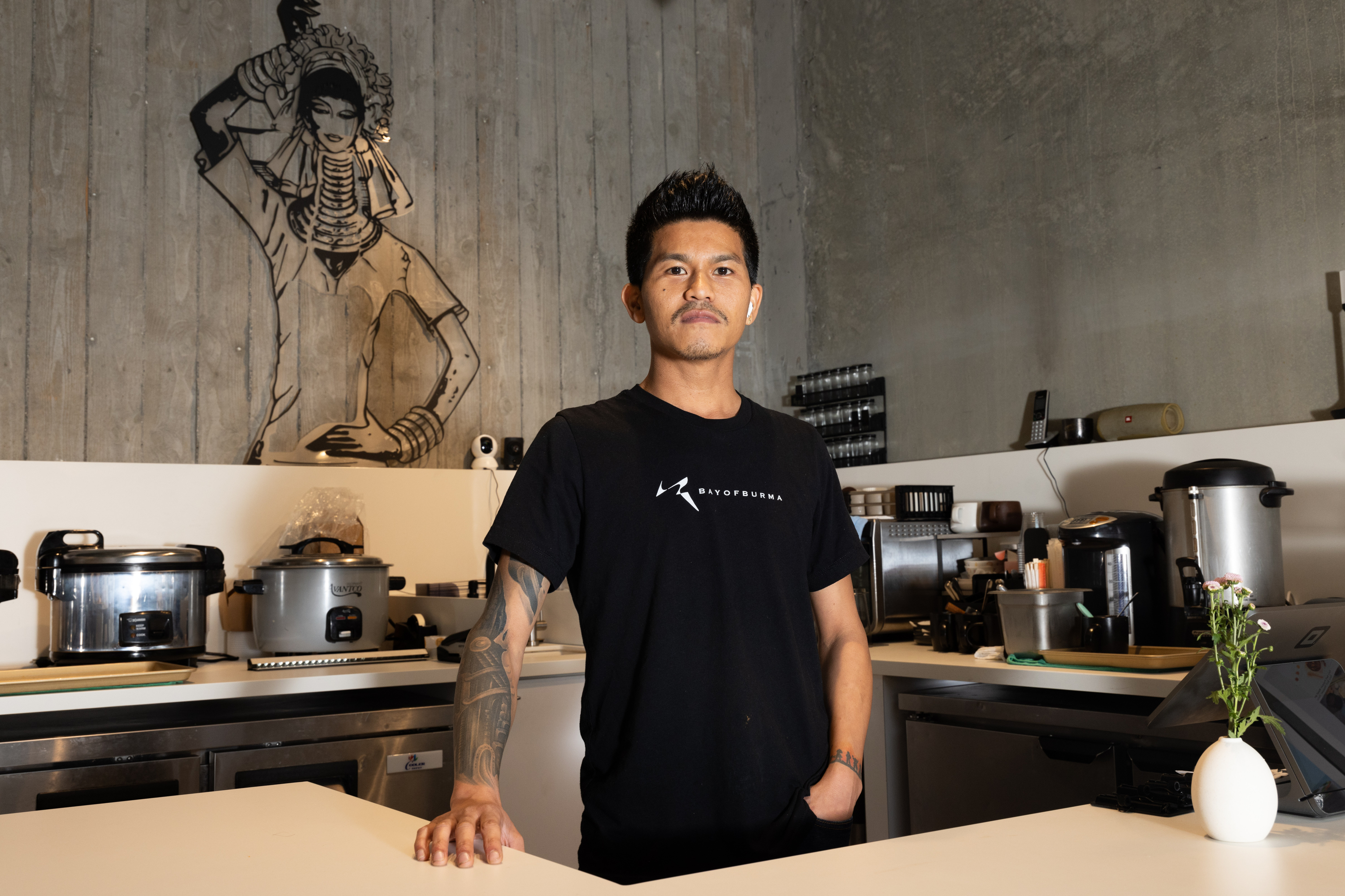 A person in a black T-shirt stands confidently in a kitchen with cooking equipment. A drawing of a woman is on the wall, and a small vase with flowers is on the counter.