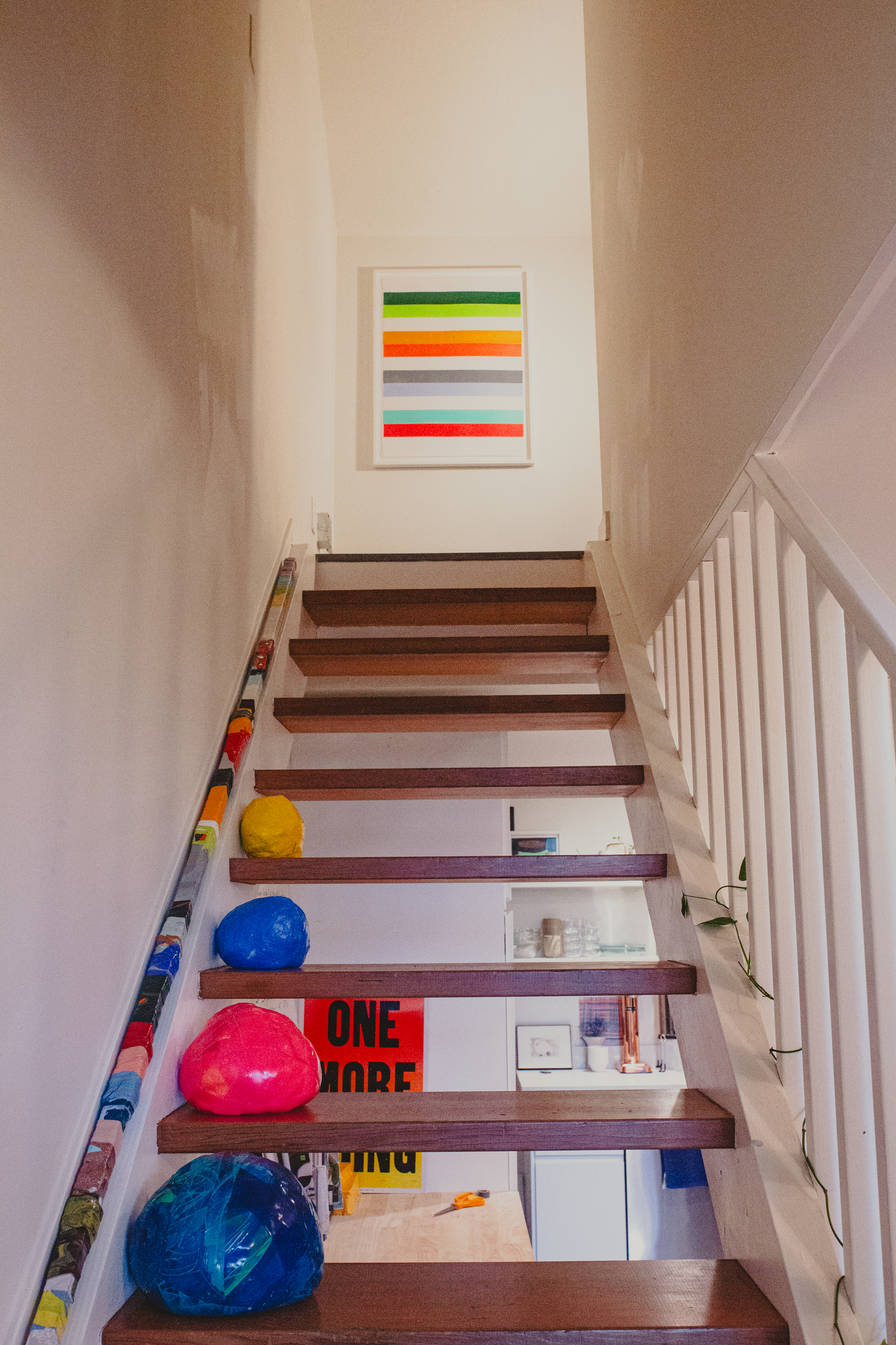 A wooden staircase decorated with colorful rocks leads up to a wall with a vibrant striped painting. The stair railing is lined with art supplies.