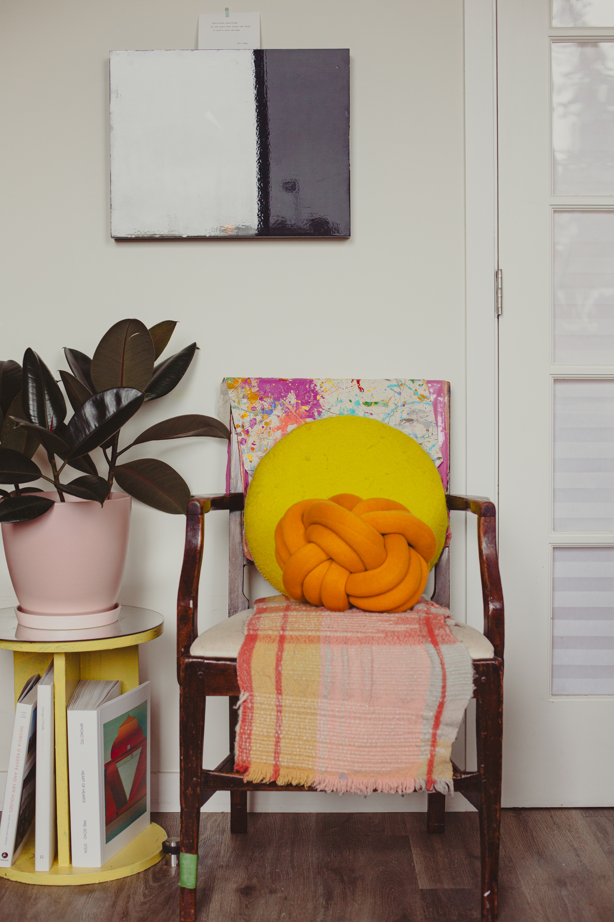 A cozy room with a wooden chair, colorful cushions, a pink potted plant on a yellow shelf with books, and abstract art on the wall.