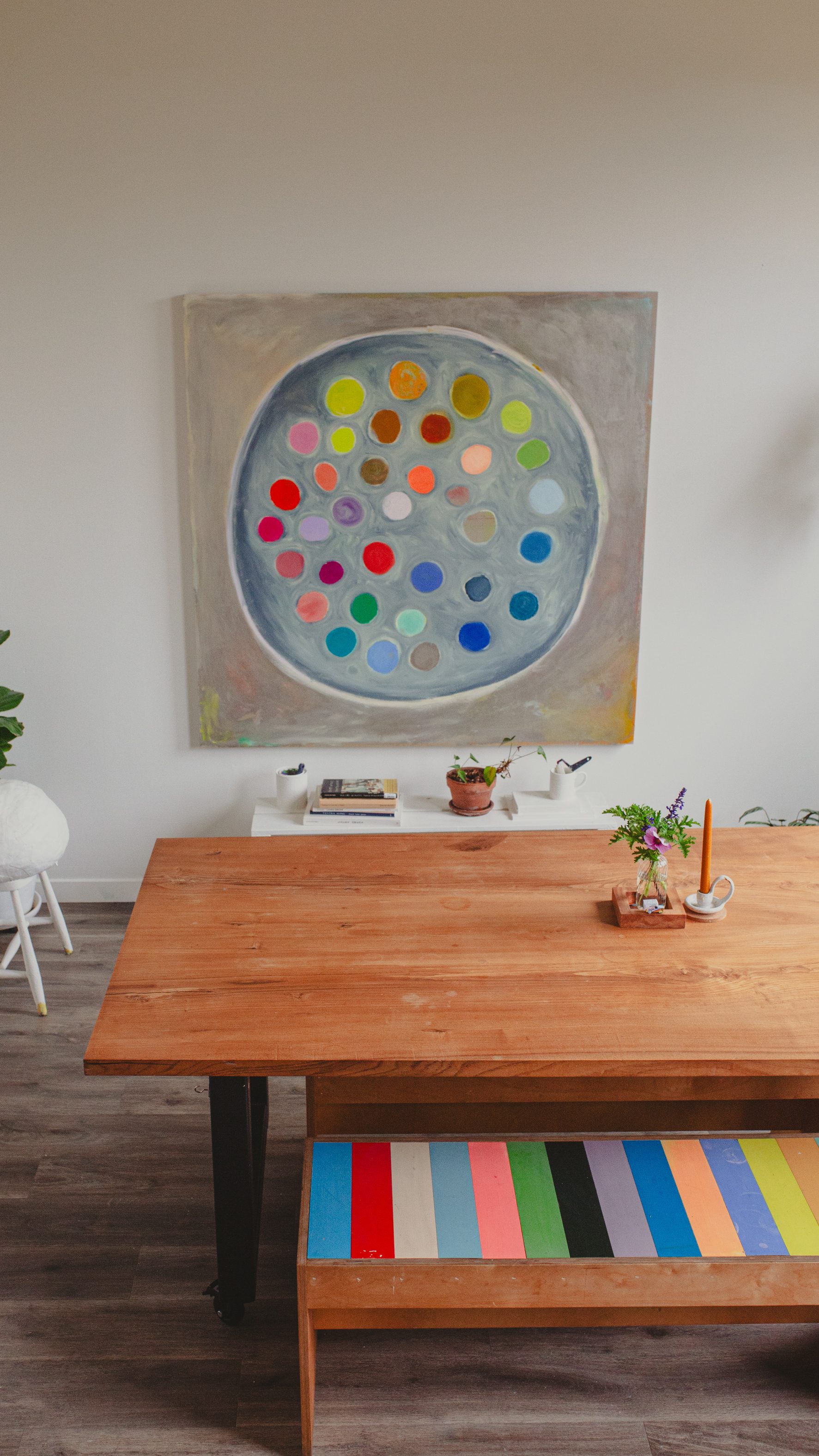 A wooden table is topped with a vase and candle. Behind it, a large colorful painting with circles hangs above a bench featuring multicolored stripes.