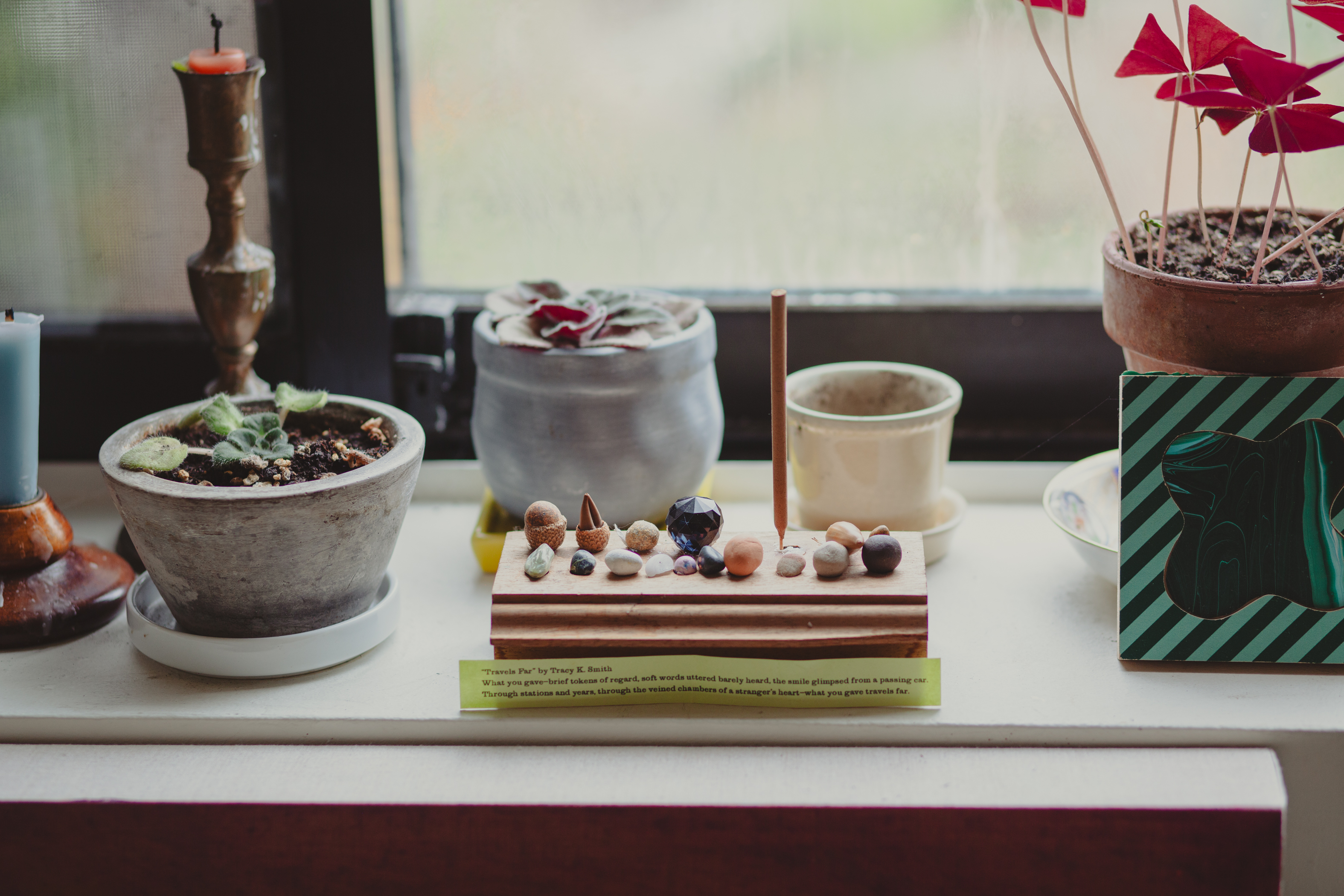 On a windowsill, there are potted plants, a candlestick, a blue candle, and a wooden tray with stones and an incense stick. Nearby rests a green-striped art piece.