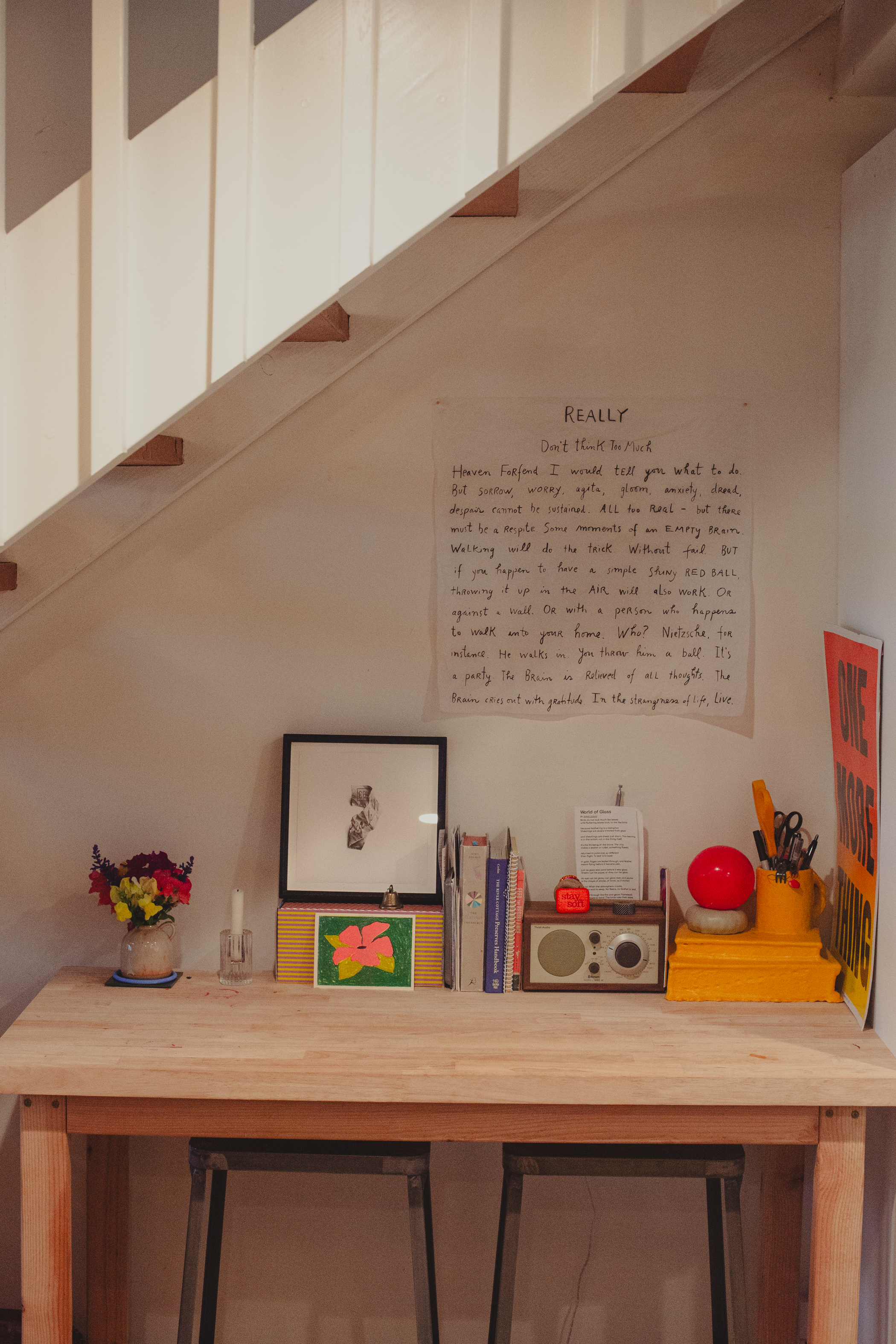 A cozy workspace under stairs features a wooden desk with books, flowers, a radio, and vibrant art. A handwritten note hangs on the wall above.