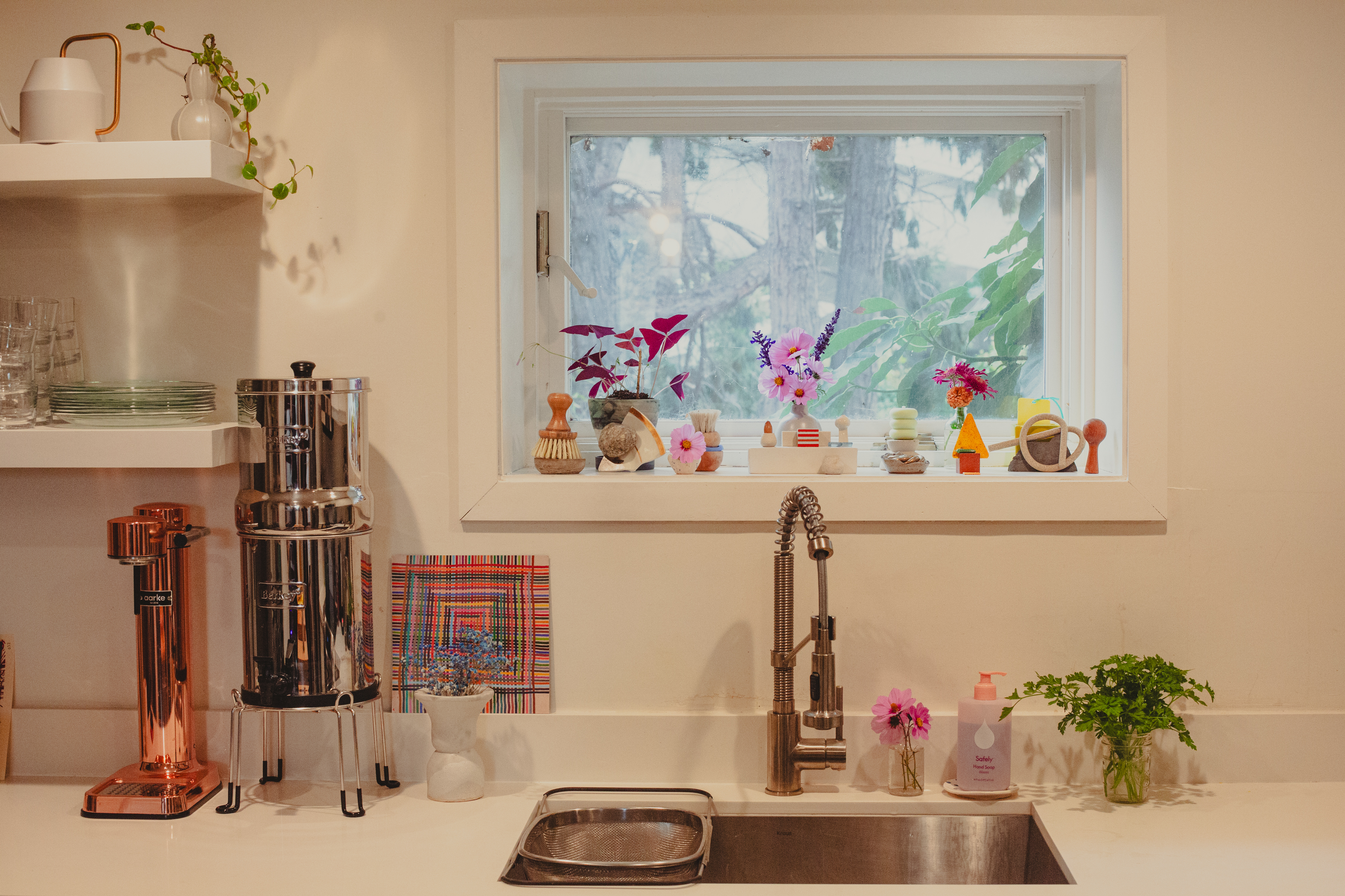 A kitchen sink with a stainless steel faucet is below a window adorned with colorful plants and decorations. Nearby are shelves with dishes and a water filter.