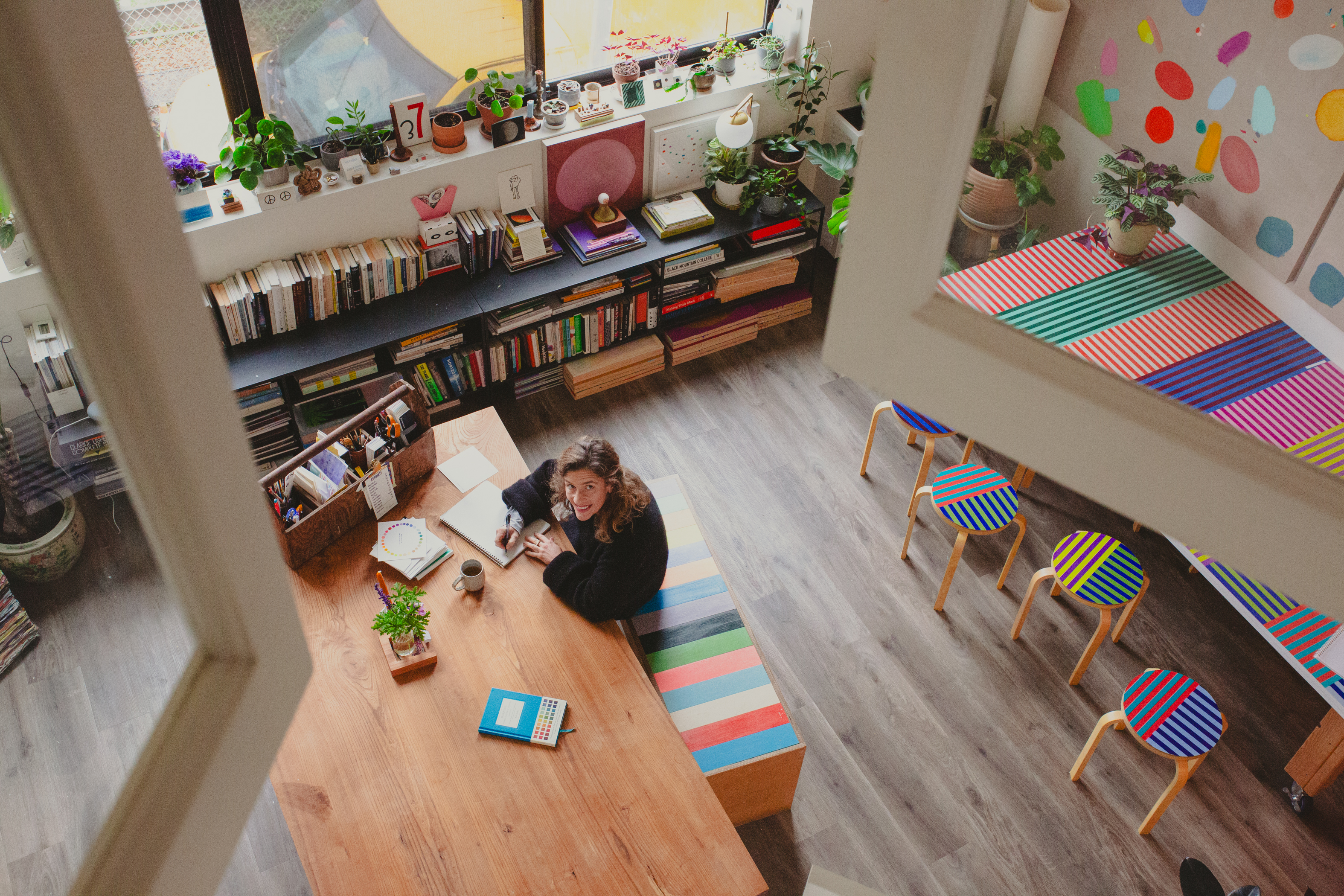 A cozy, colorful home office with a wooden table, bookshelves, and plants. A person writes in a notebook, surrounded by colorful patterns and art supplies.