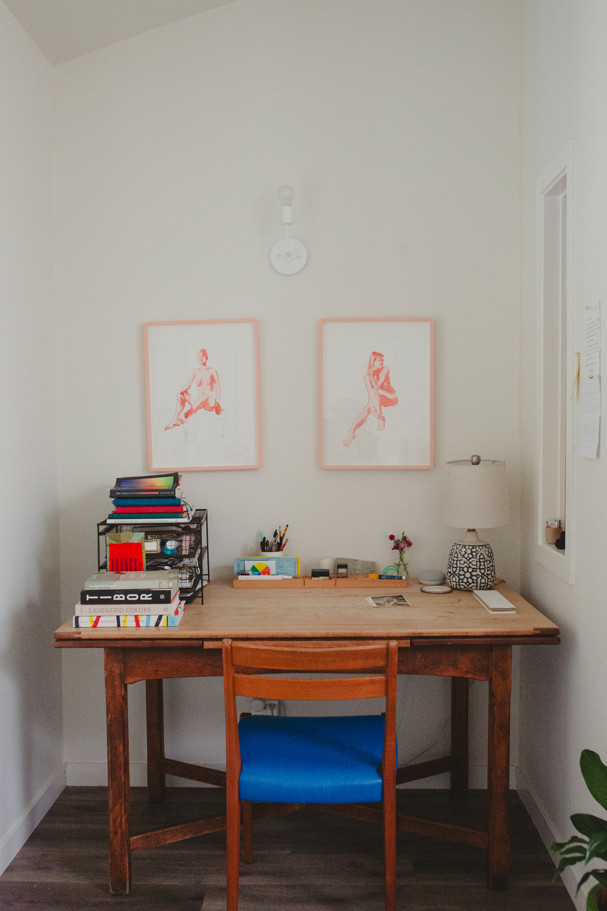 A tidy wooden desk with a blue chair is adorned with books, a lamp, stationery, and two framed red artworks on the wall. A plant peeks from the side.