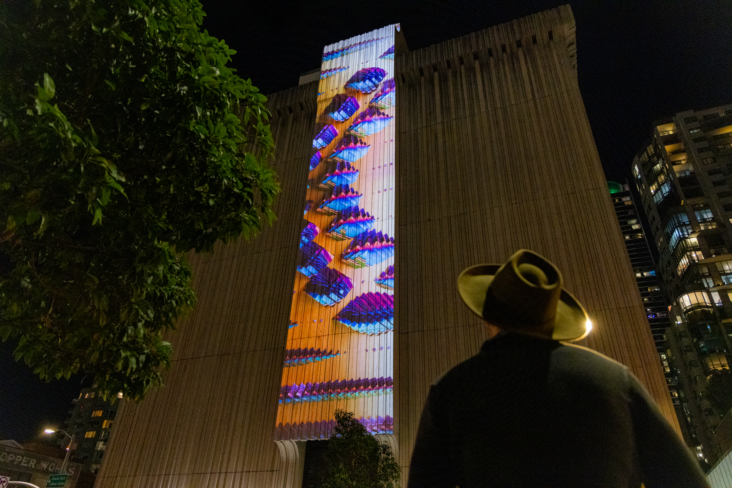 A person in a hat looks up at a tall building with vibrant, colorful projections at night, featuring abstract patterns. A tree and buildings surround the scene.