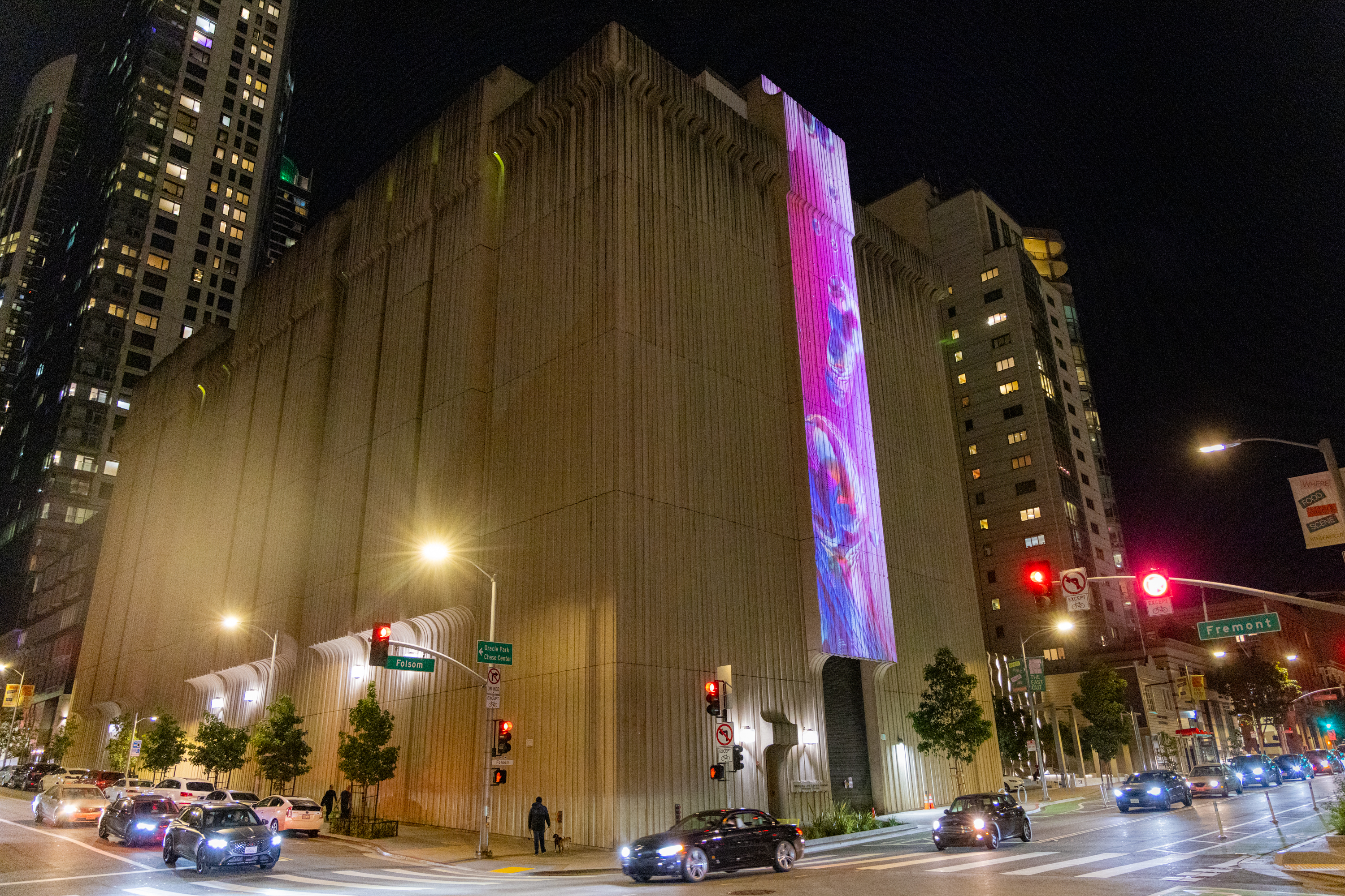 A large building with textured walls is illuminated at night. A colorful digital display shows abstract patterns on the facade. Cars and traffic lights are visible.