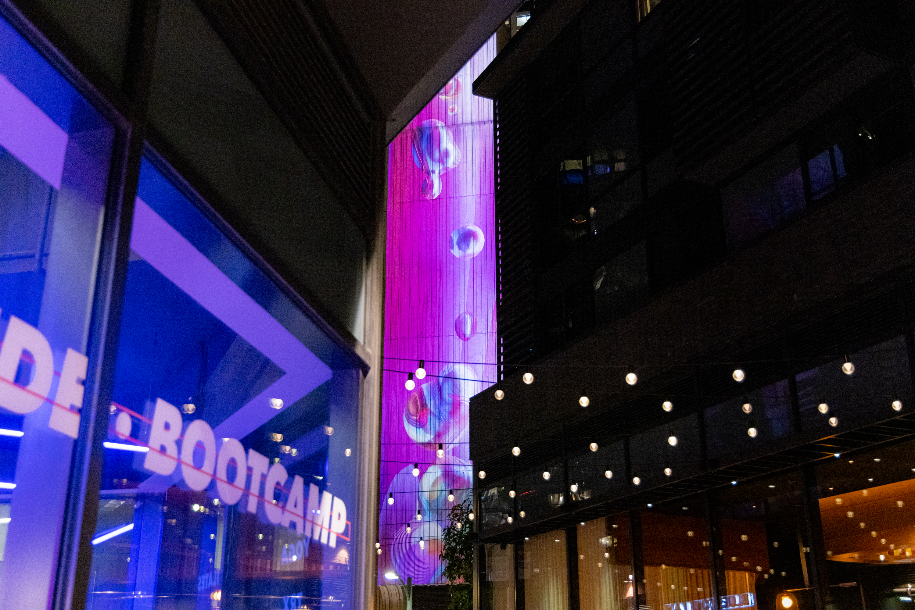 The image shows a vibrant city scene with a pink-lit building displaying bubbles, a &quot;BOOTCAMP&quot; sign in blue light, and string lights overhead.