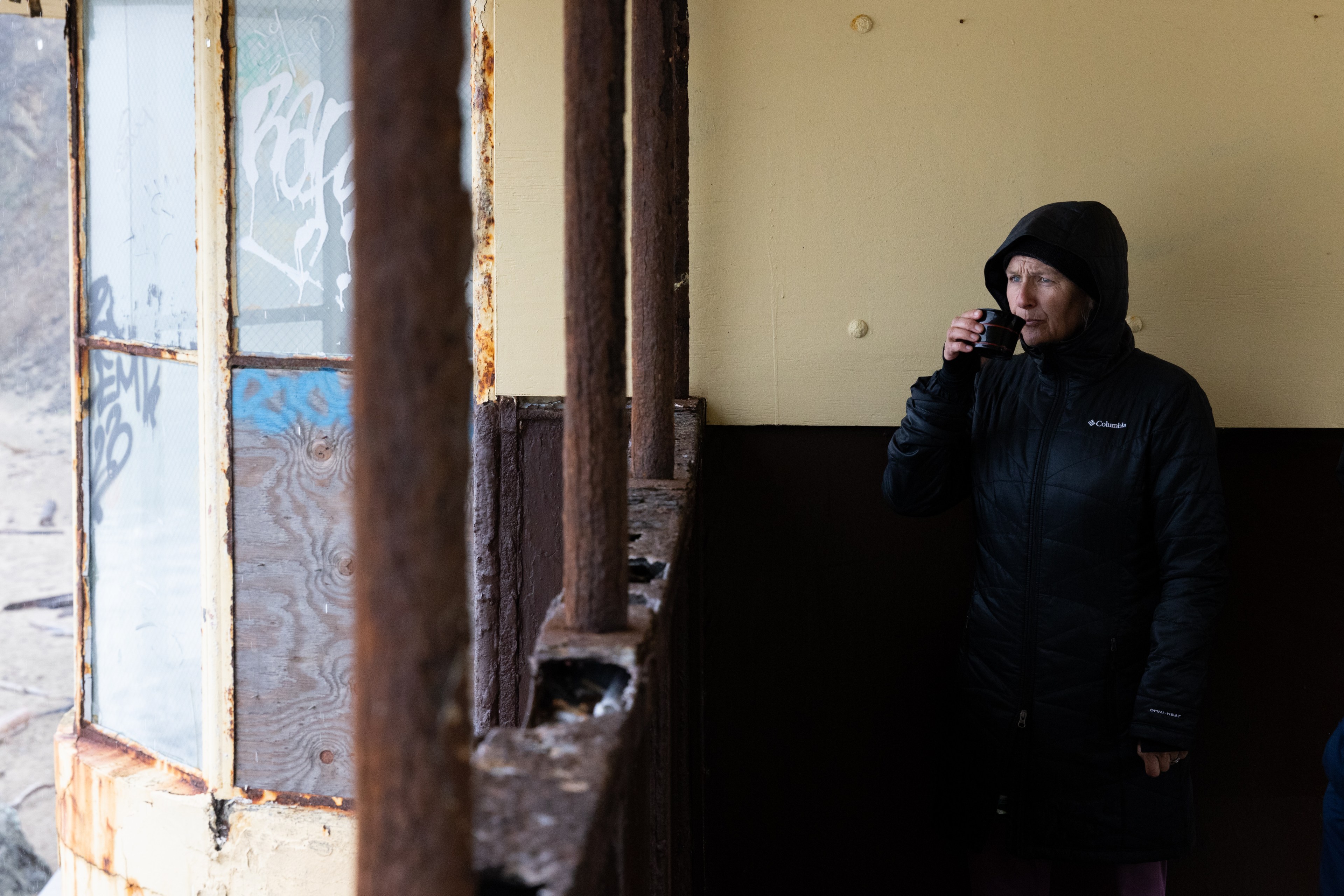 A person in a black hooded coat drinks from a cup in a rusted, graffiti-marked structure with weathered windows. The ambiance is cold and gritty.