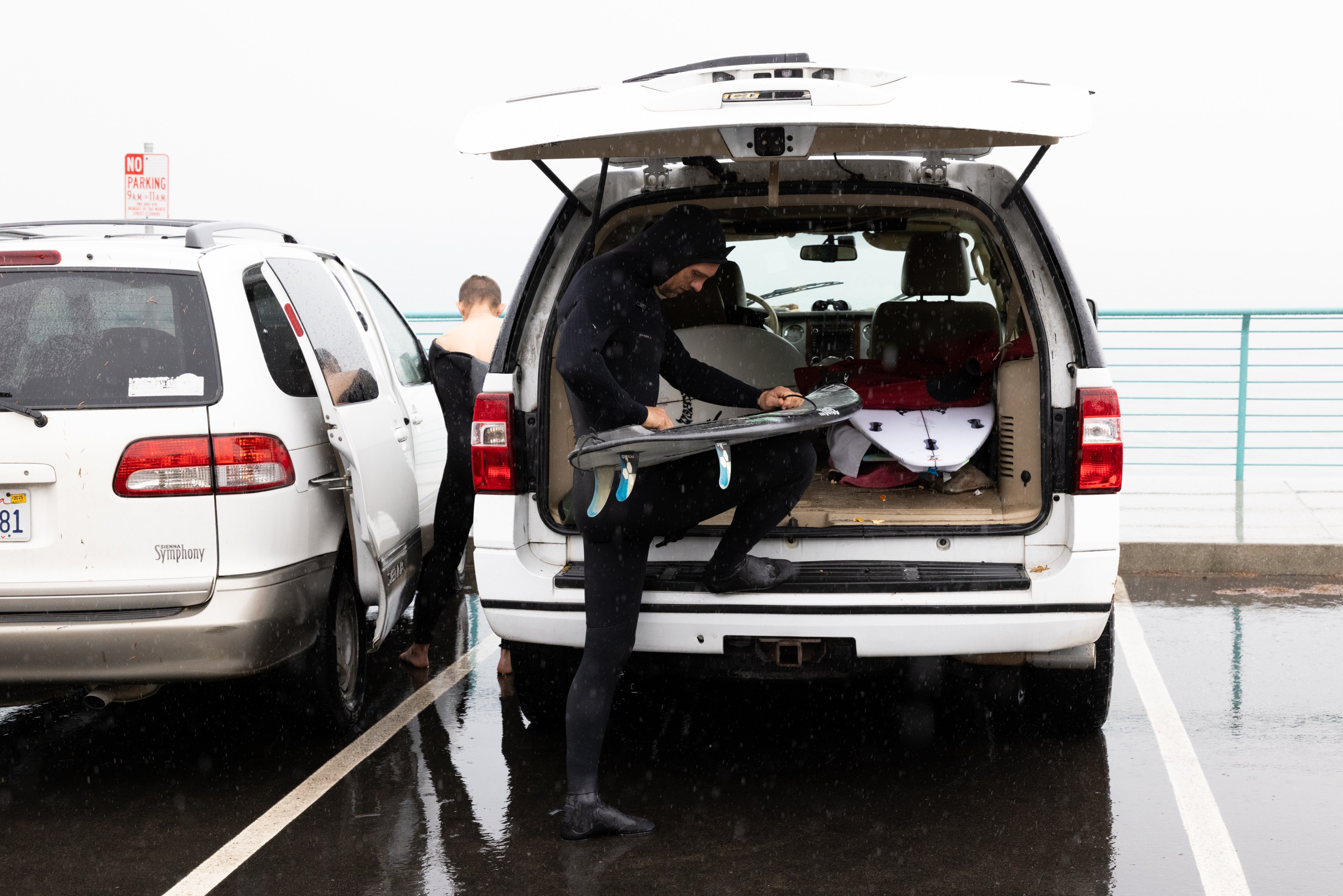 Two people in wetsuits prepare surfboards at the open back of an SUV in a rainy parking lot, with wet ground and a &quot;No Parking&quot; sign visible.