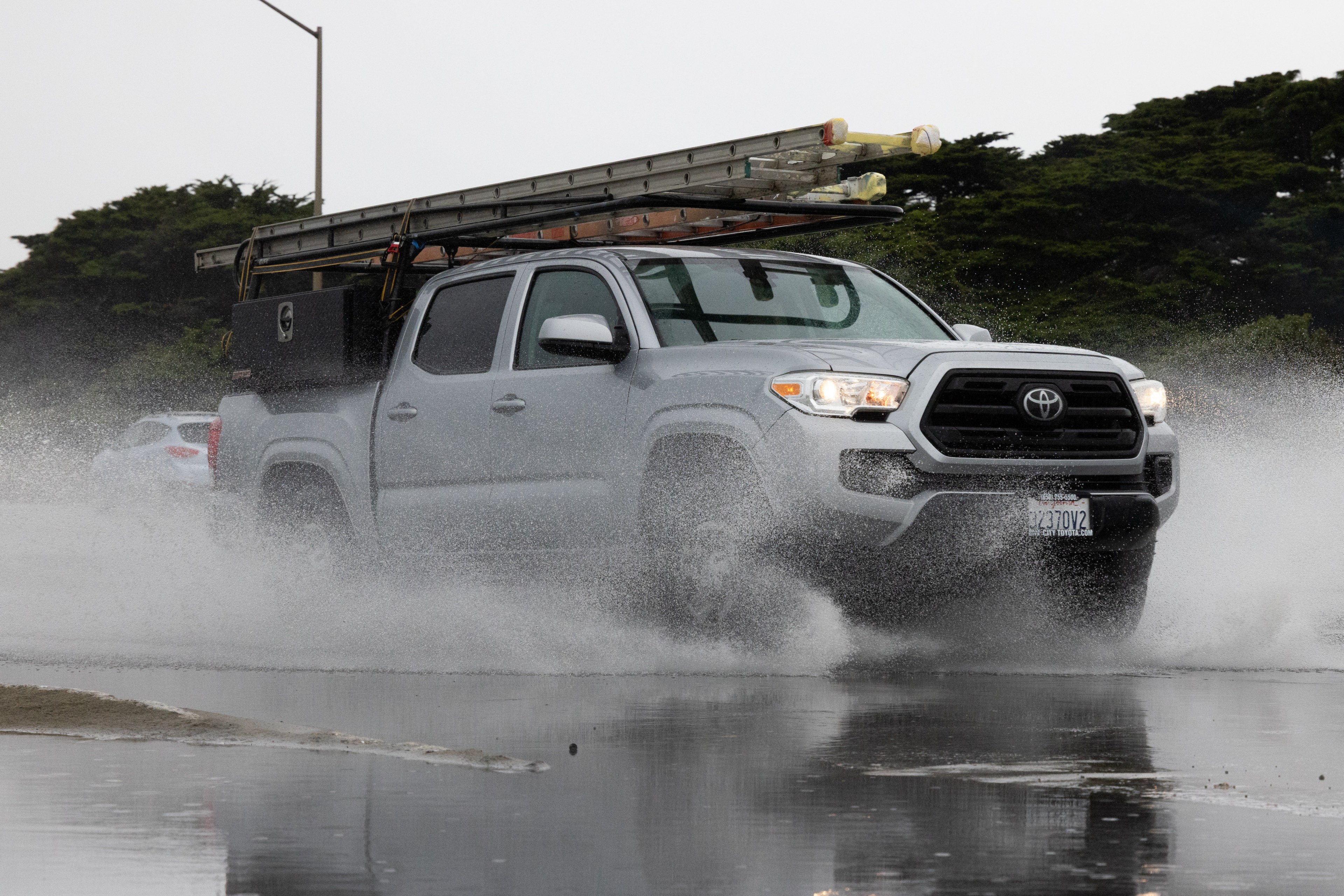 A gray truck drives through a large puddle, splashing water. It has ladders secured on top and a dark toolbox in the back, with trees and a streetlamp in the background.