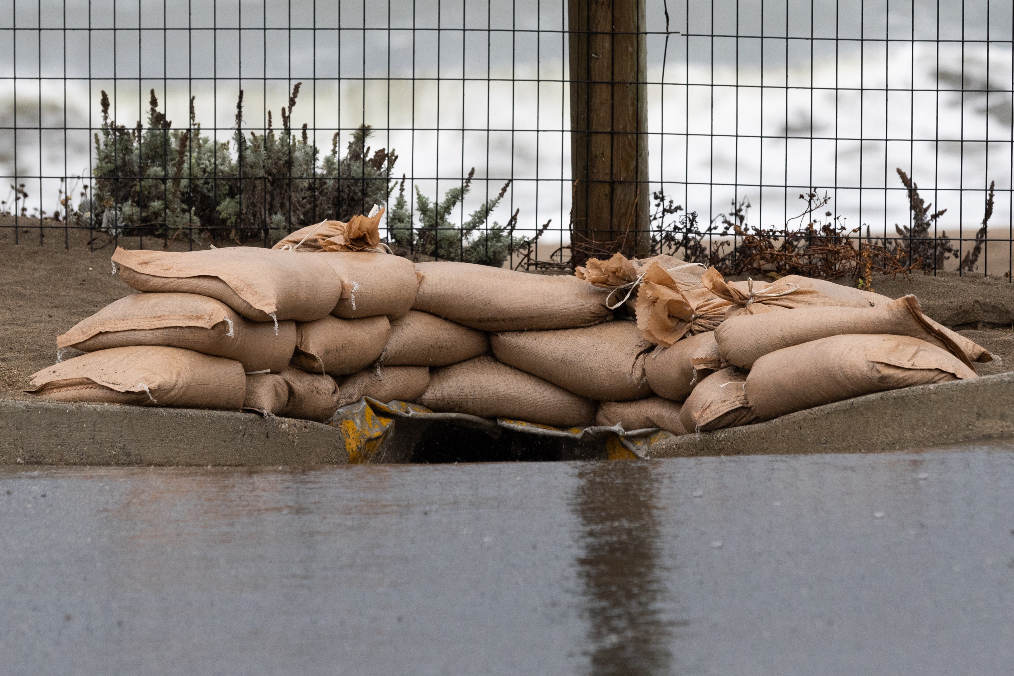 A row of sandbags is stacked in front of a drainage pipe. A wire fence and some foliage are in the background, with a hint of an overcast sky.