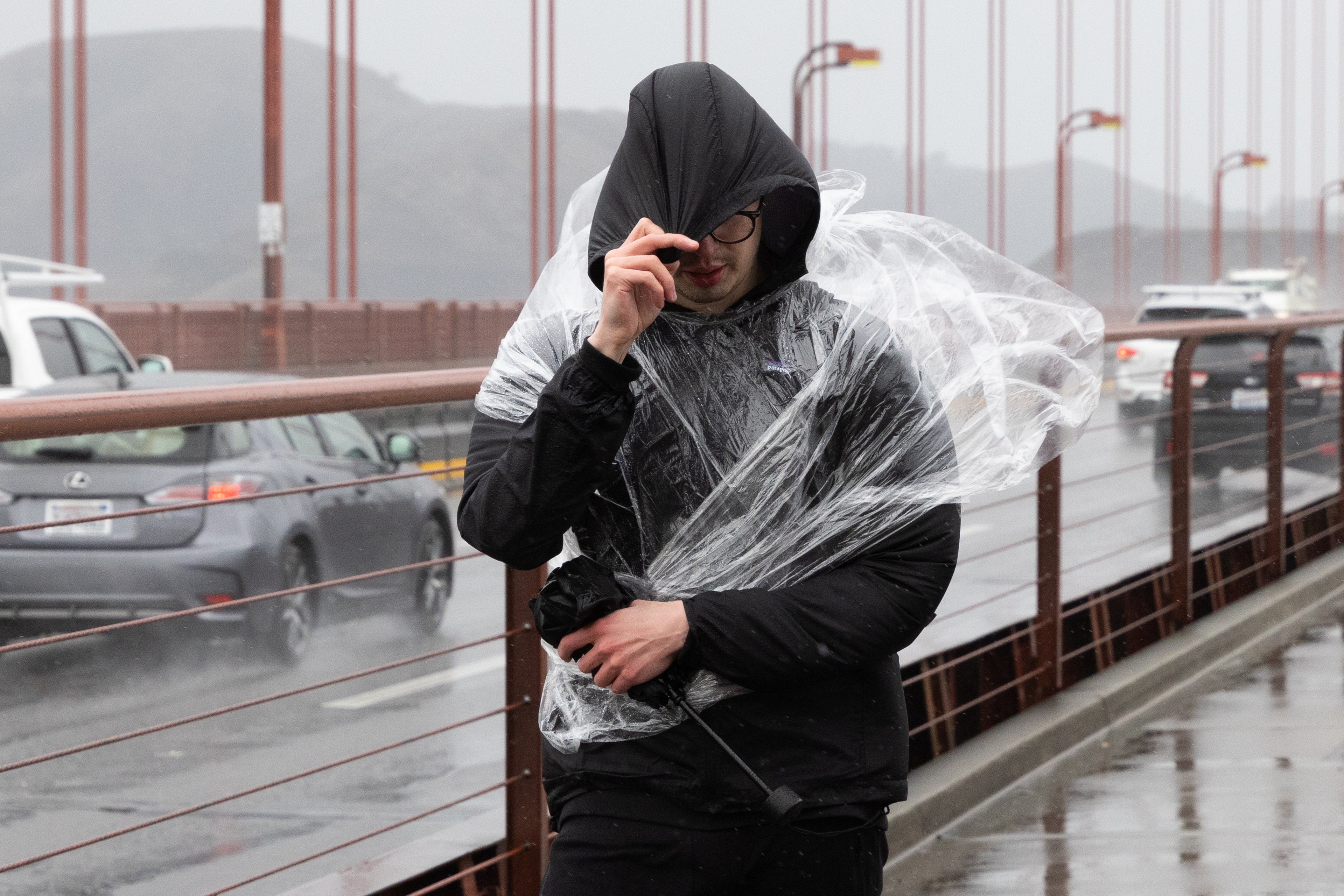 A person in a black jacket with a hood walks across a rainy bridge. They hold a plastic sheet wrapped around them while adjusting their glasses. Cars drive by in the background.