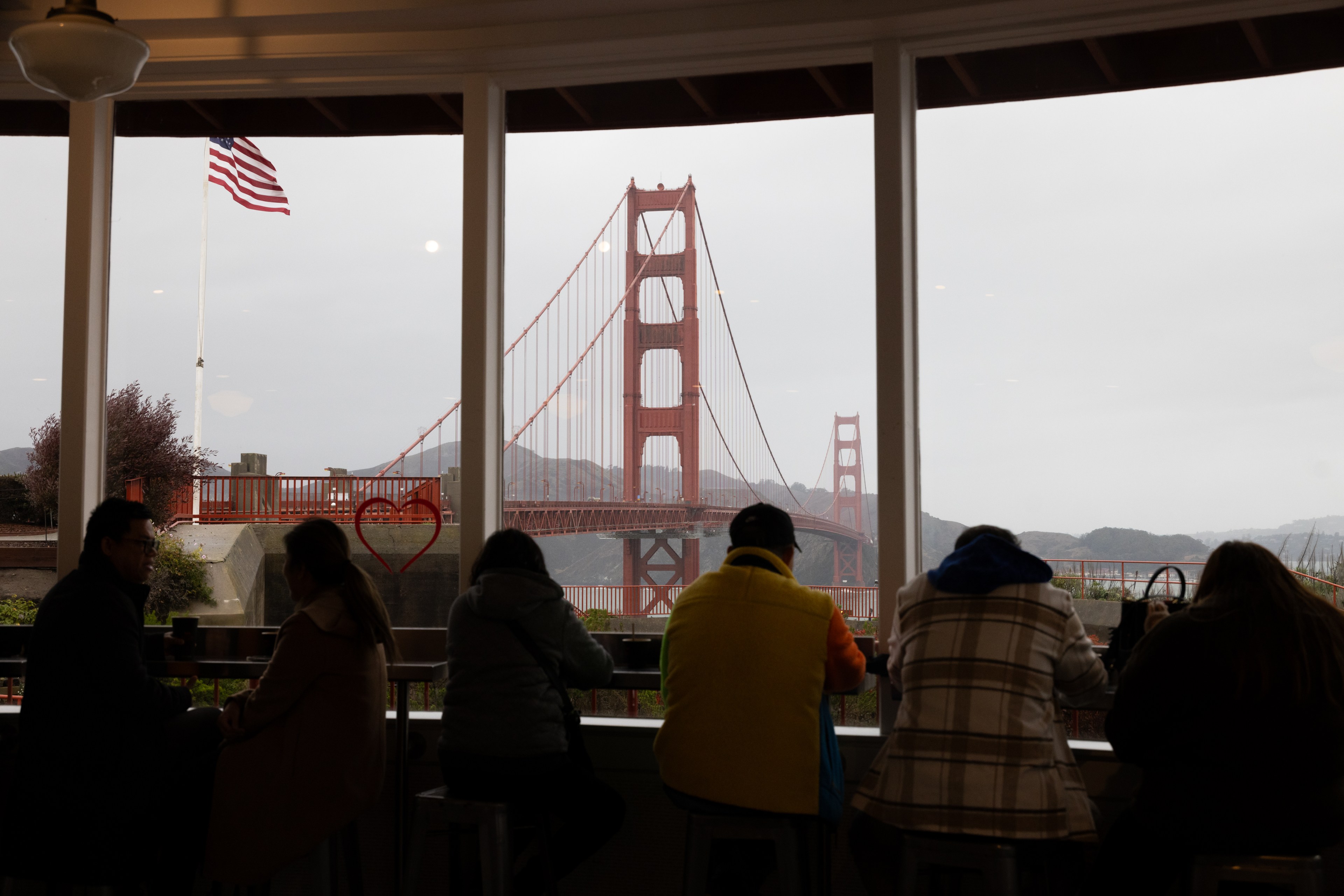 People sit inside a café, silhouetted against large windows framing the Golden Gate Bridge and an American flag outside, with overcast skies.