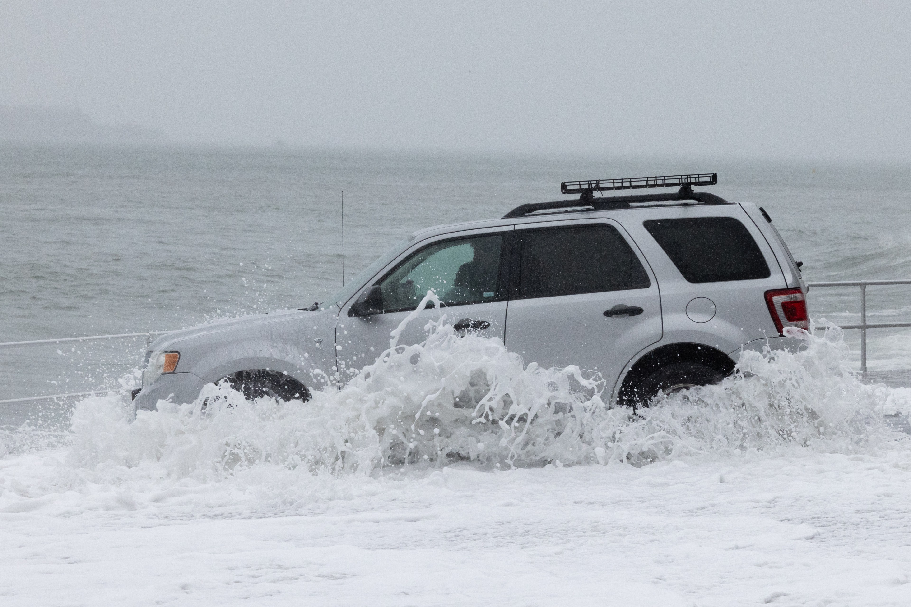 A silver SUV drives through water splashing against its sides, near a coastal area. The ocean is visible in the background, and the sky is overcast.