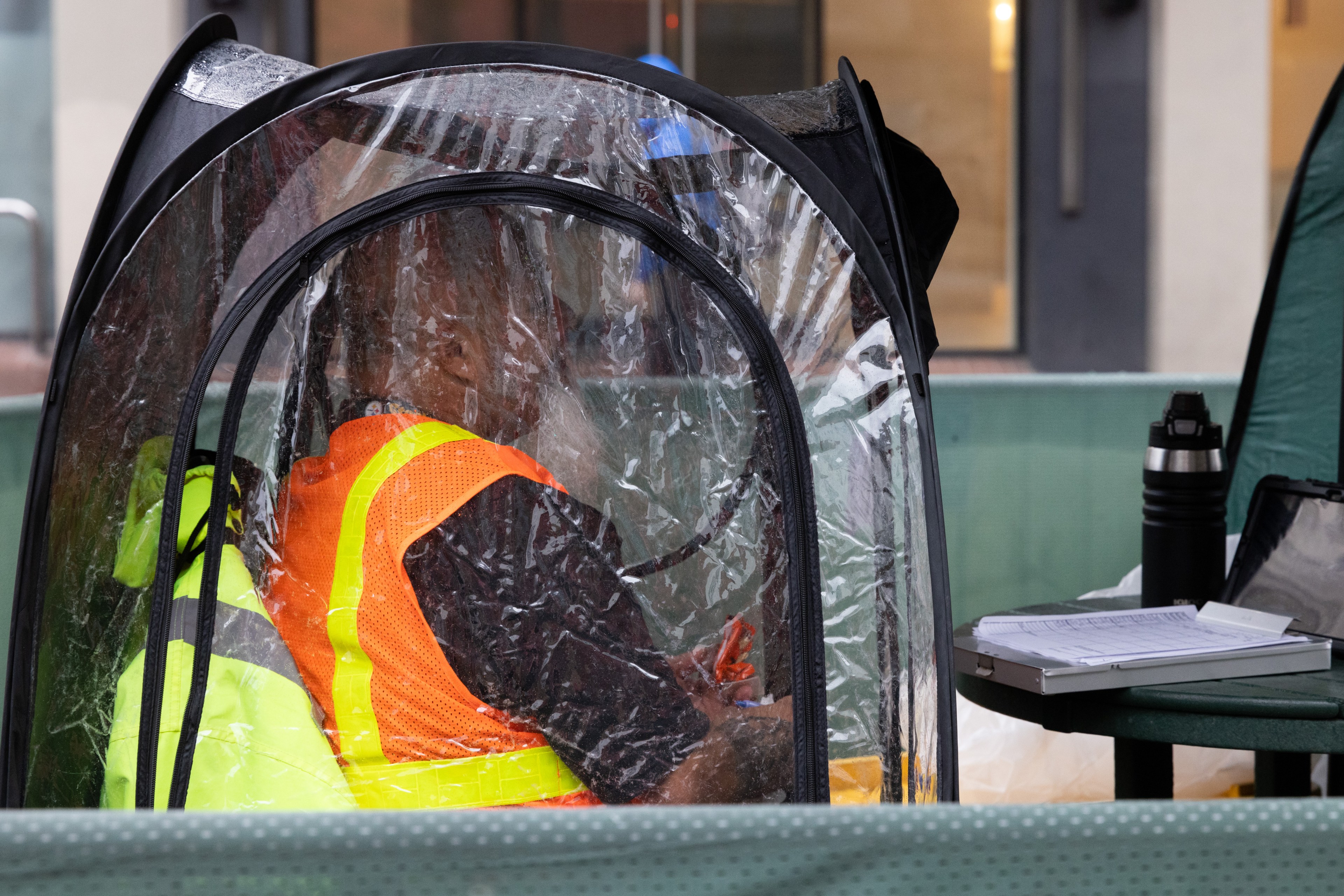 A person in a neon vest is seated inside a small transparent tent. Beside them is a table with a thermos, a notebook, and a digital device.