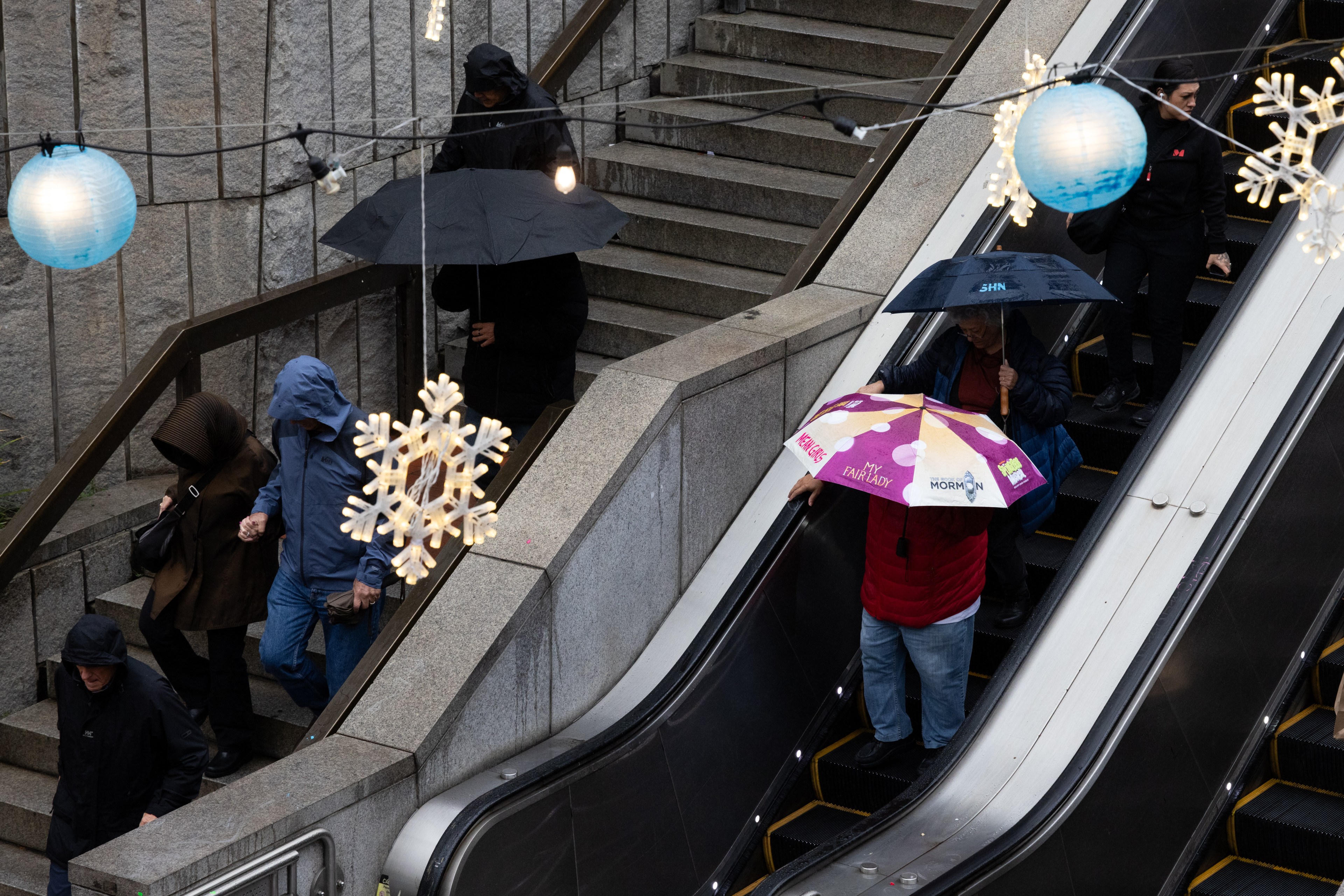 People with umbrellas are using an escalator and stairs on a rainy day. Decorative lights, including snowflakes and blue globes, hang overhead.