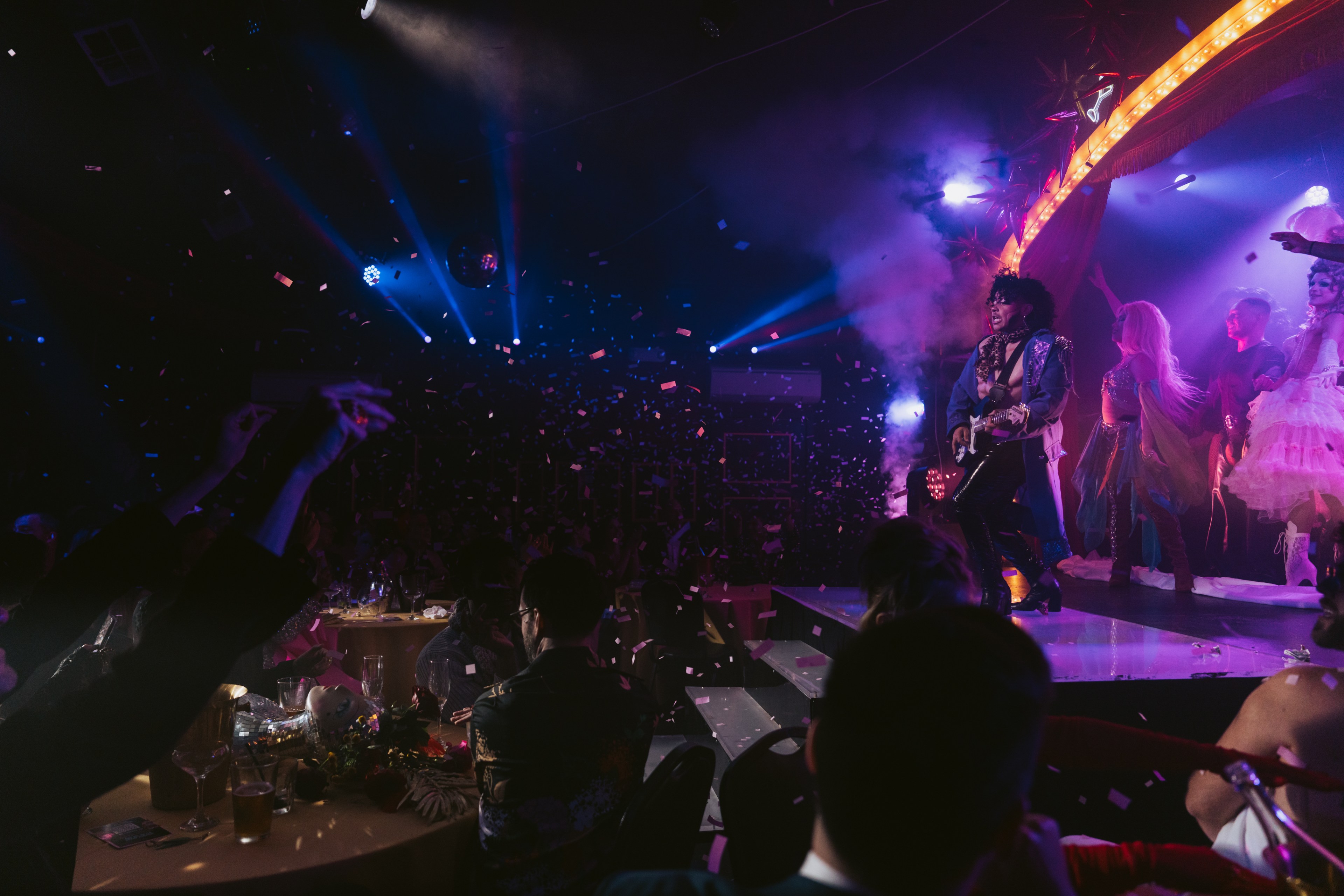 A lively stage performance features a costumed musician playing guitar, surrounded by colorful lights, confetti, and an engaged audience.