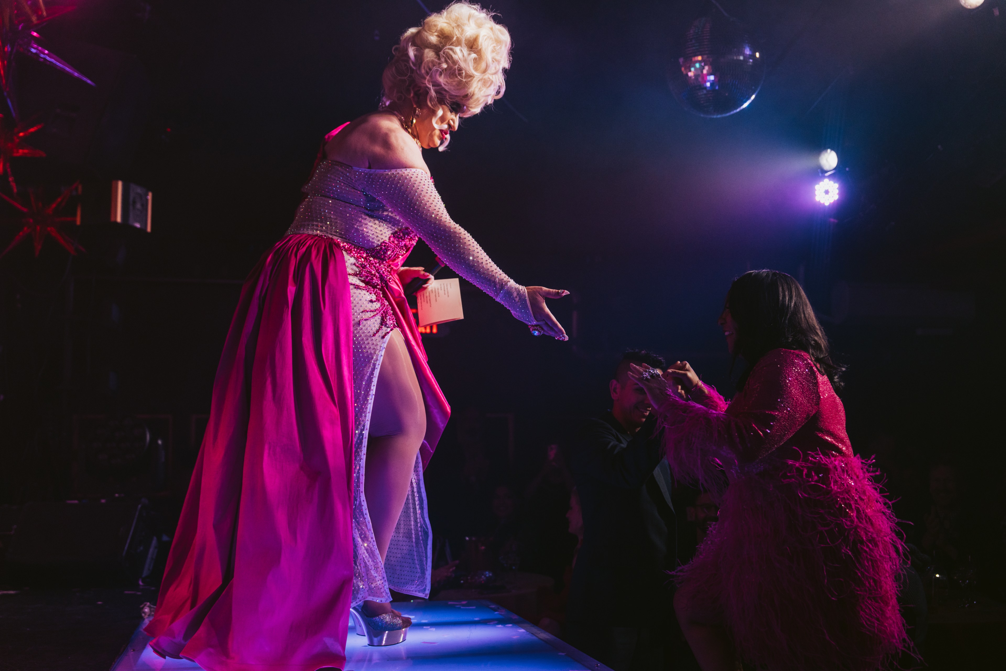 A performer in a sparkling pink and silver dress with a voluminous wig reaches toward an audience member. The scene is brightly lit with a disco ball overhead.