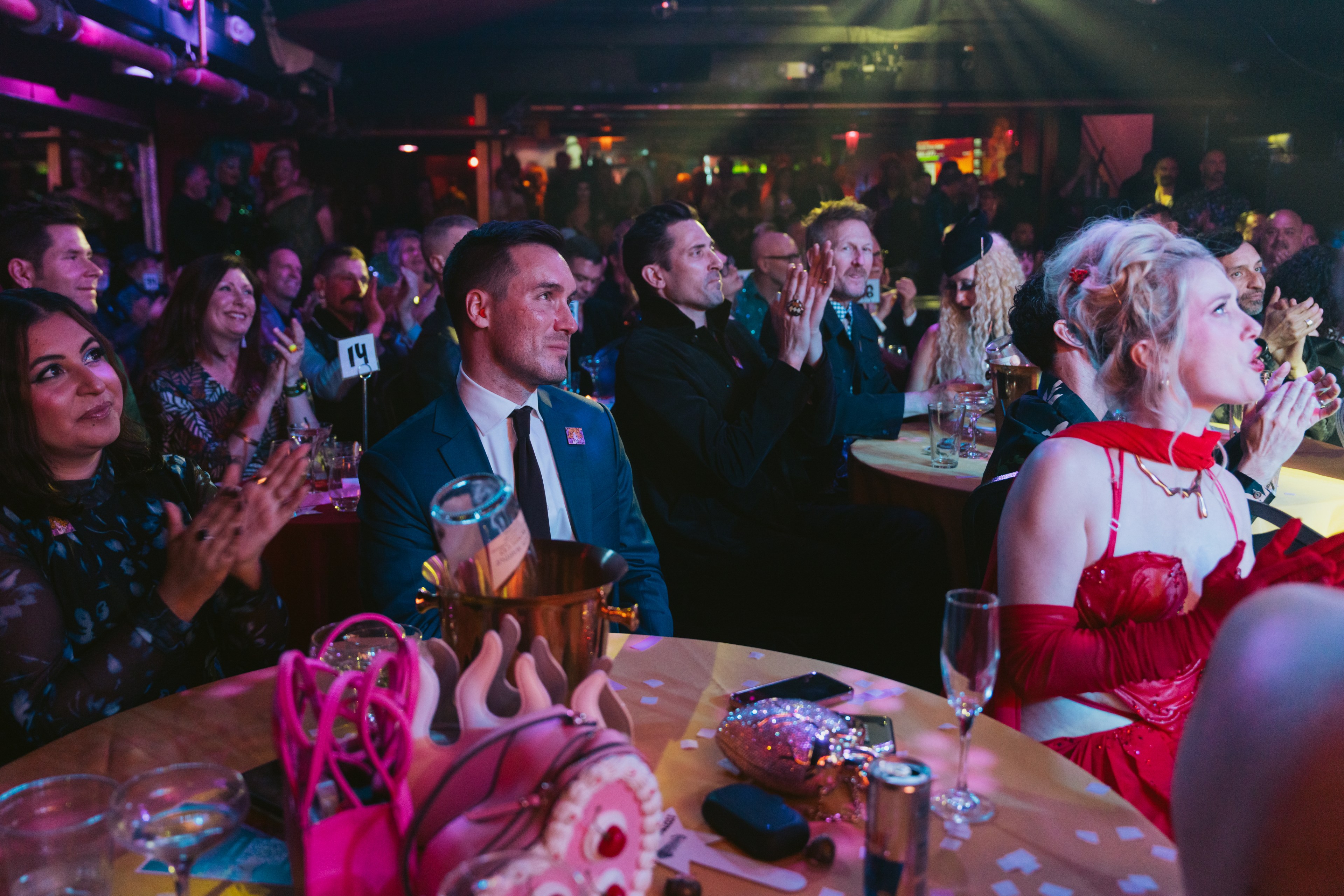 A lively audience sits at tables in a dimly lit venue, clapping and watching attentively. One woman in a vibrant red outfit is prominently visible at the front.