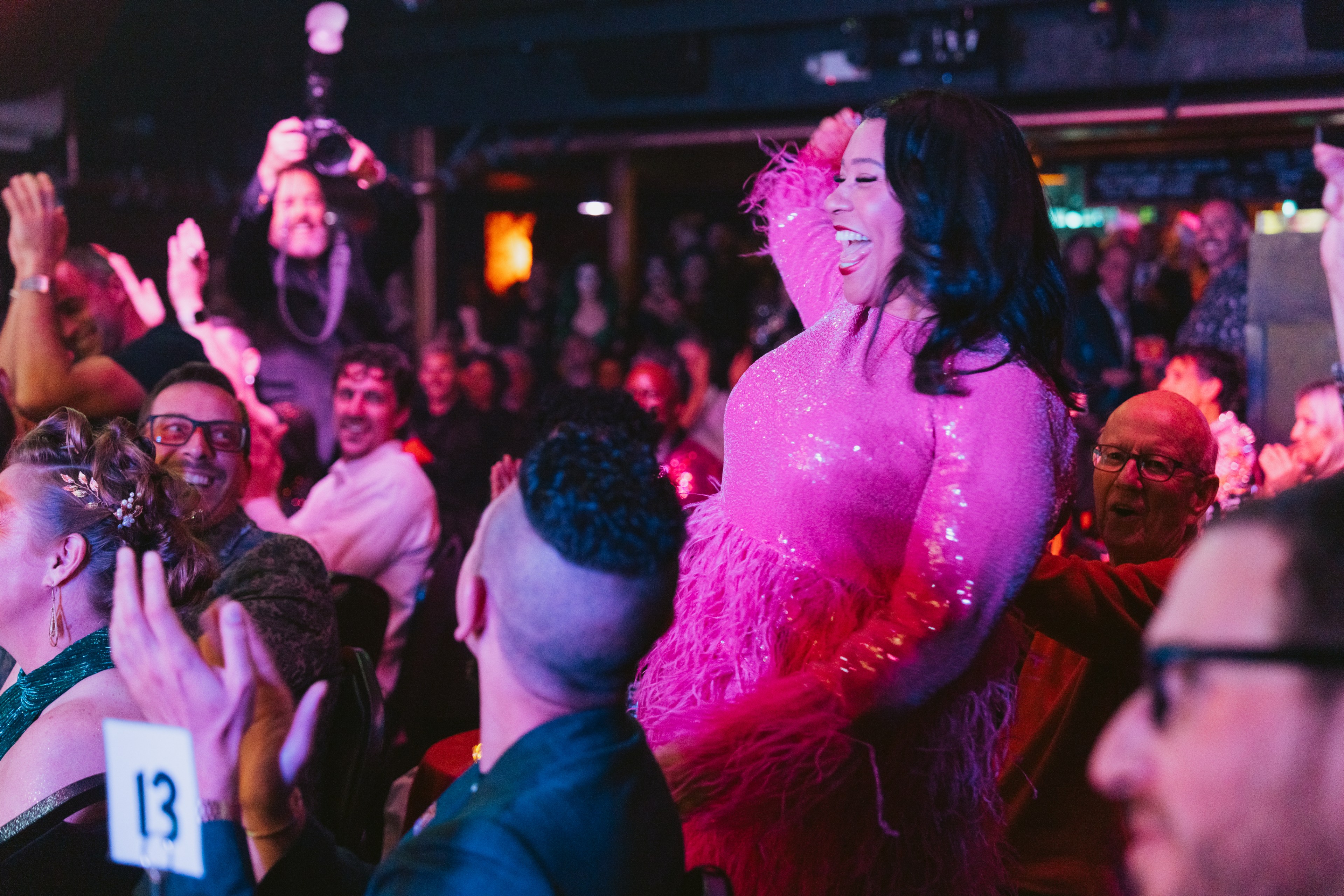 A lively scene shows a woman in a sparkling pink dress with feathers, smiling and engaging with a cheering audience in a dimly lit venue.