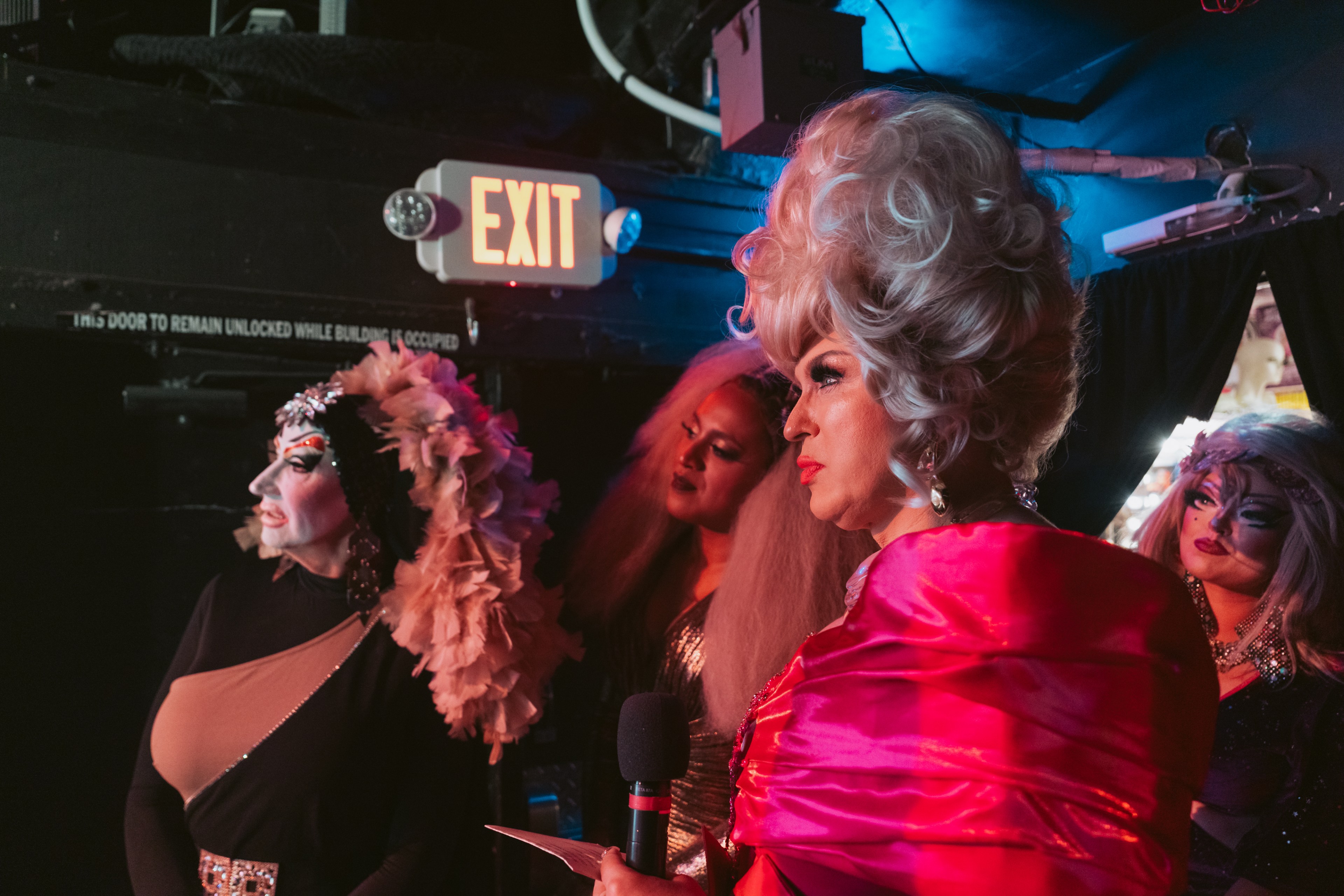 Four people in glamorous costumes and dramatic makeup stand backstage under an &quot;EXIT&quot; sign, one holding a microphone, all looking focused.