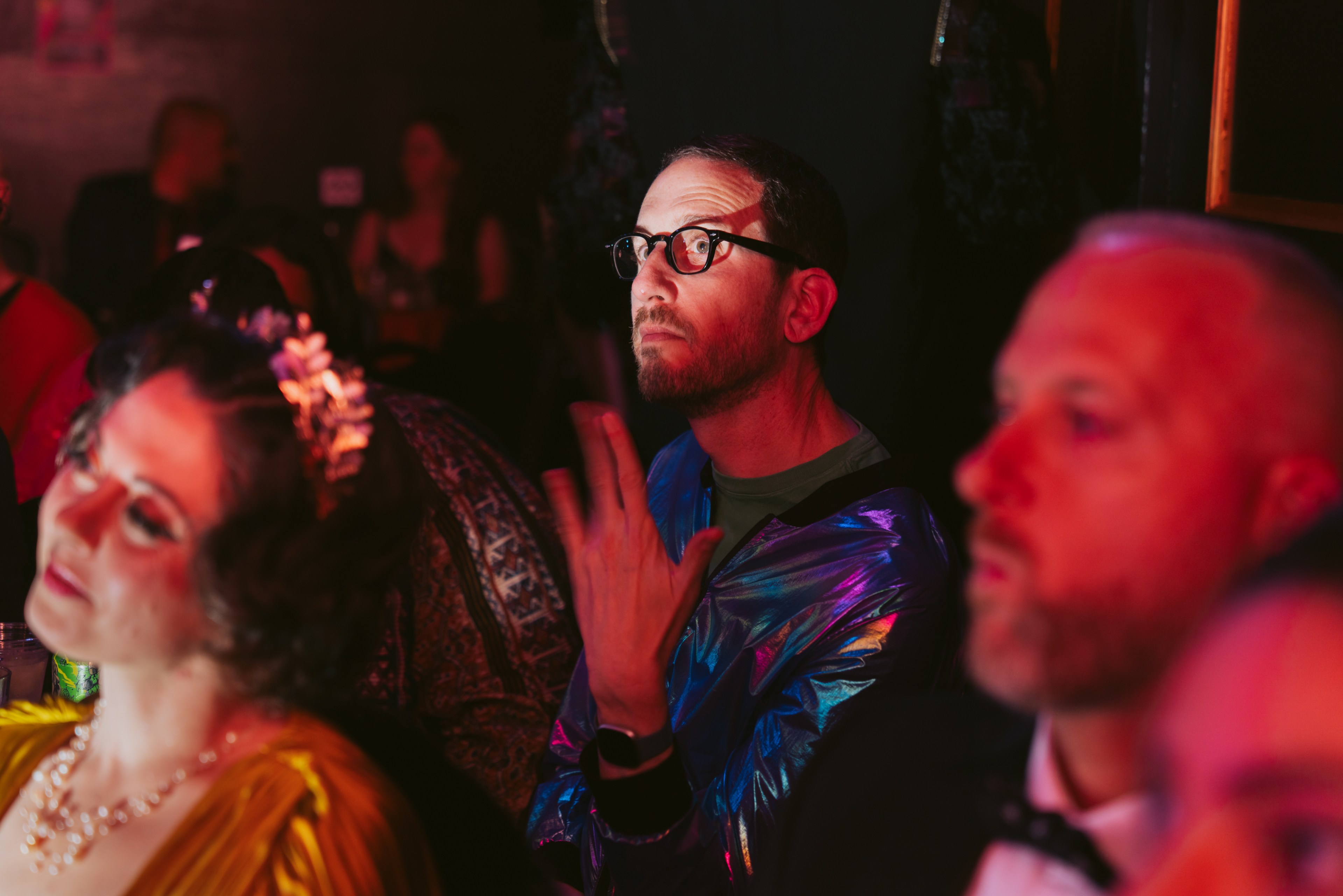 A man in glasses wearing a shiny blue jacket looks upwards thoughtfully in a dimly lit room, surrounded by people in colorful attire.