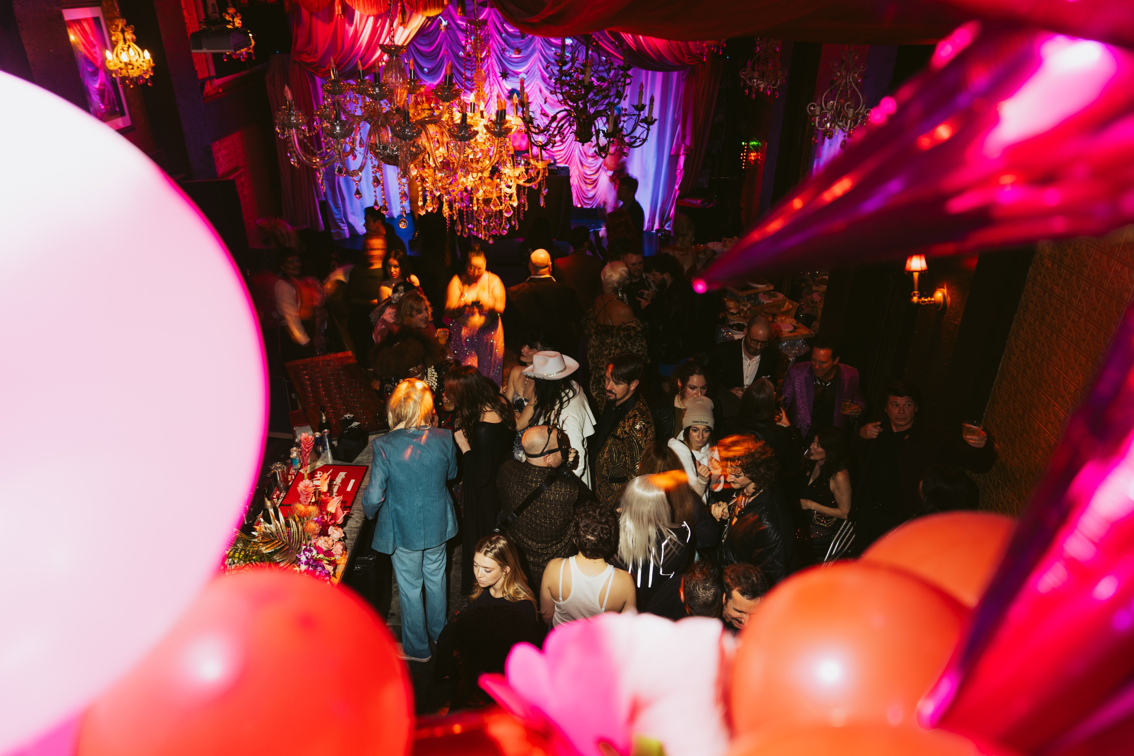 A lively party with a crowd under ornate chandeliers and vibrant lights. People are mingling, some in costumes, and balloons add a festive touch.