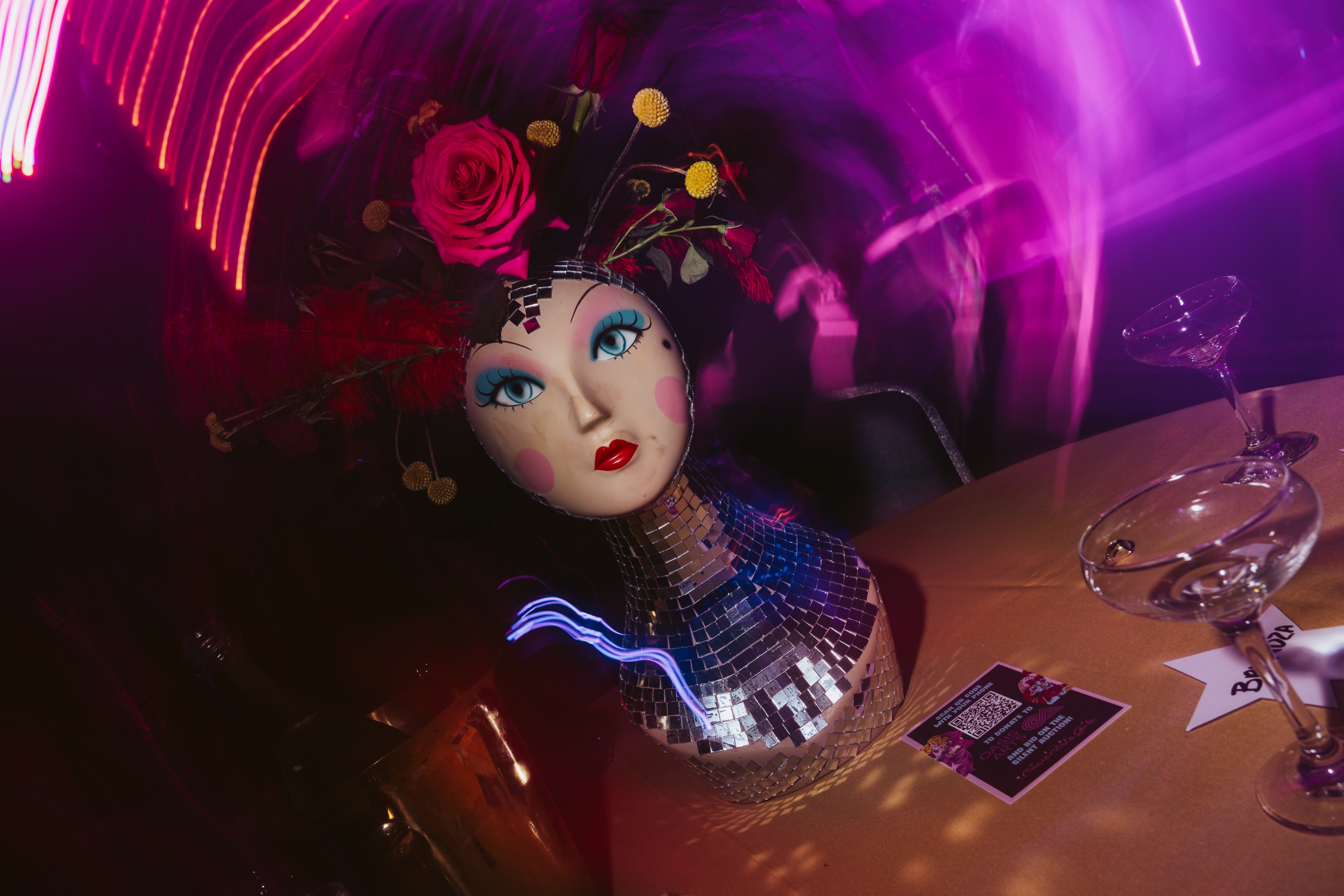 A mannequin head with blue eyes and red lips is adorned with flowers and feathers atop a mirrored base. It's placed on a table with cocktail glasses and a flyer, bathed in pink lighting.