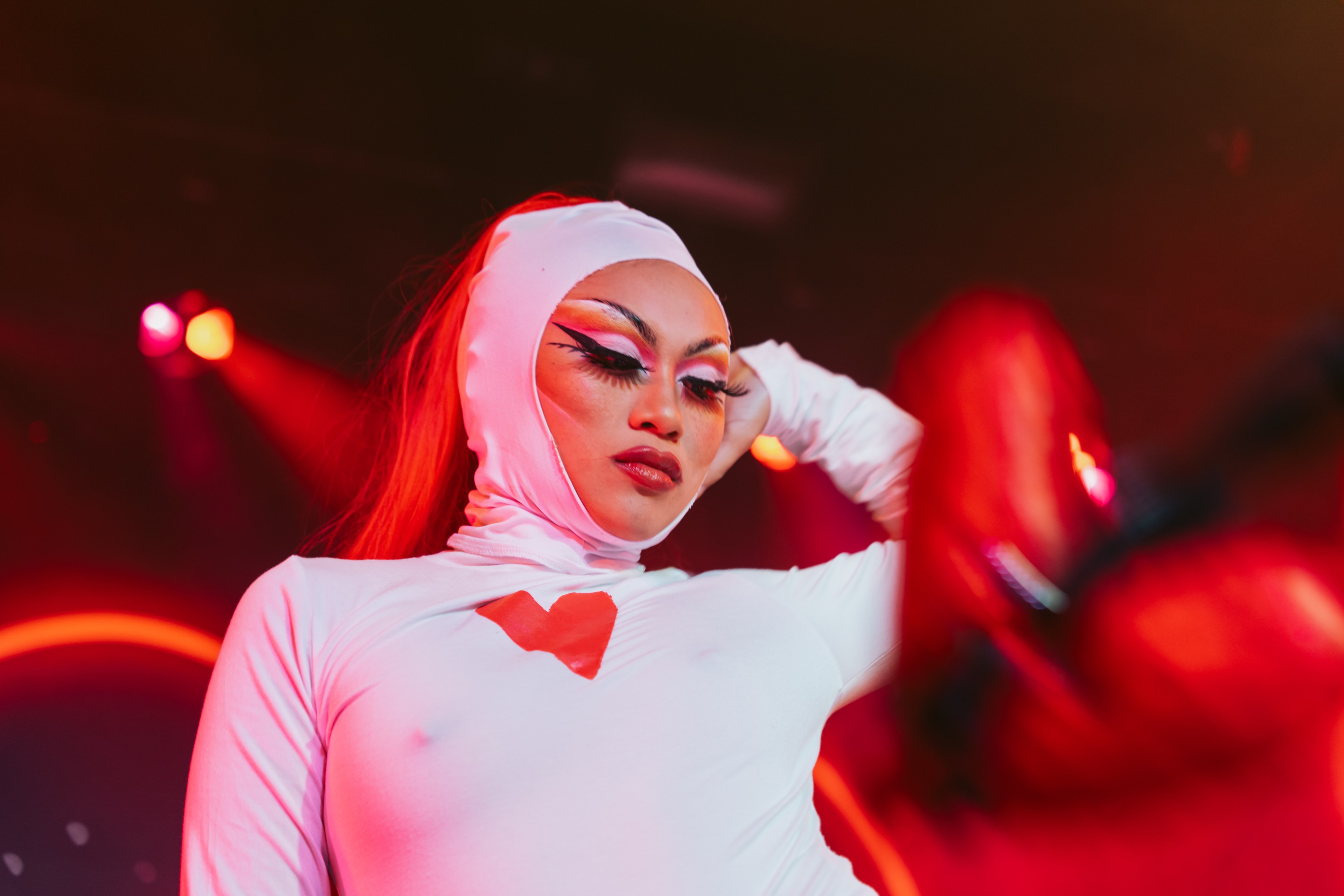 A performer in a white outfit with a heart design poses under red stage lights, with dramatic makeup and their eyes closed.