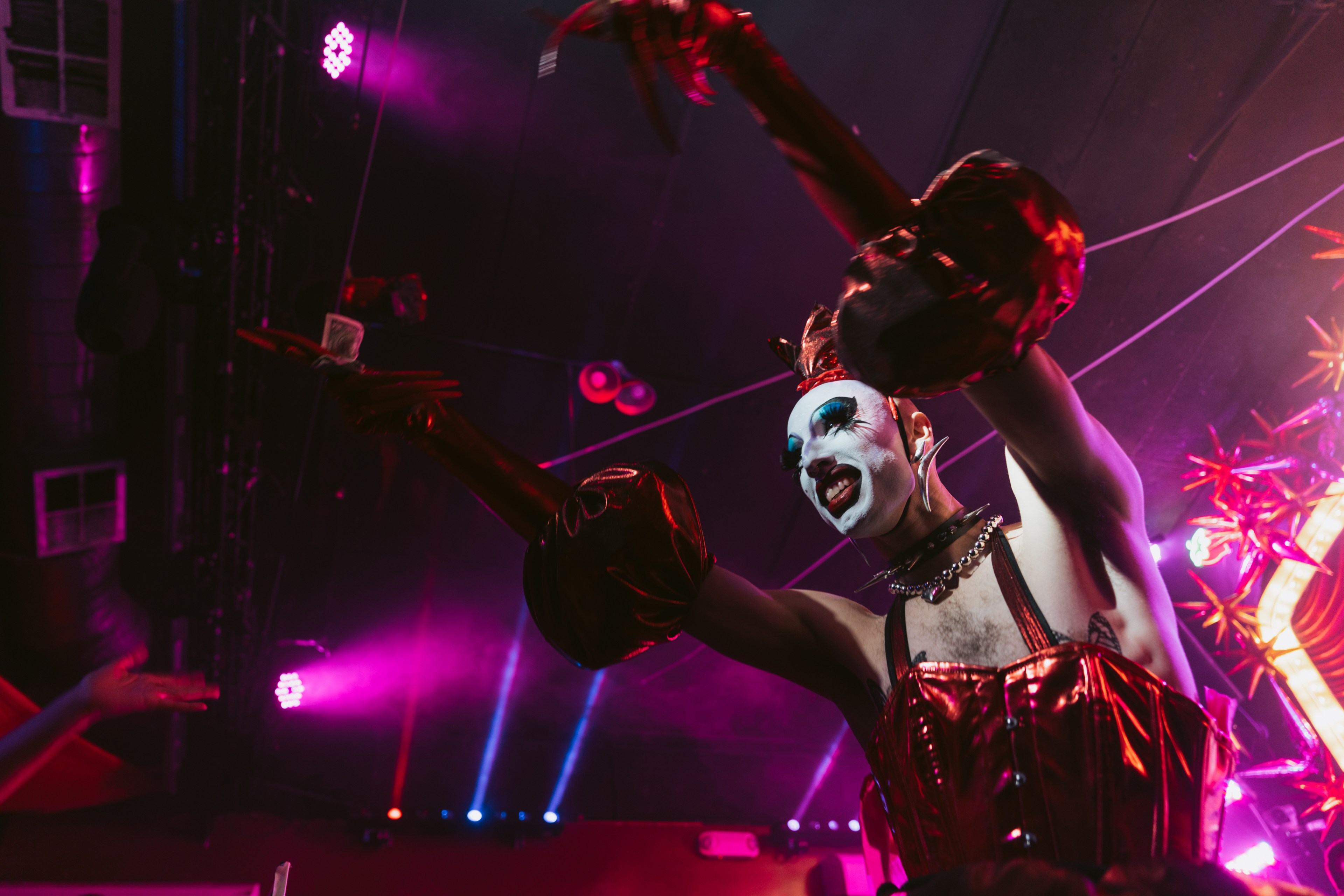 A performer in dramatic makeup and a shiny red outfit gesticulates on stage under vibrant pink and purple lights, holding a card between long fingers.