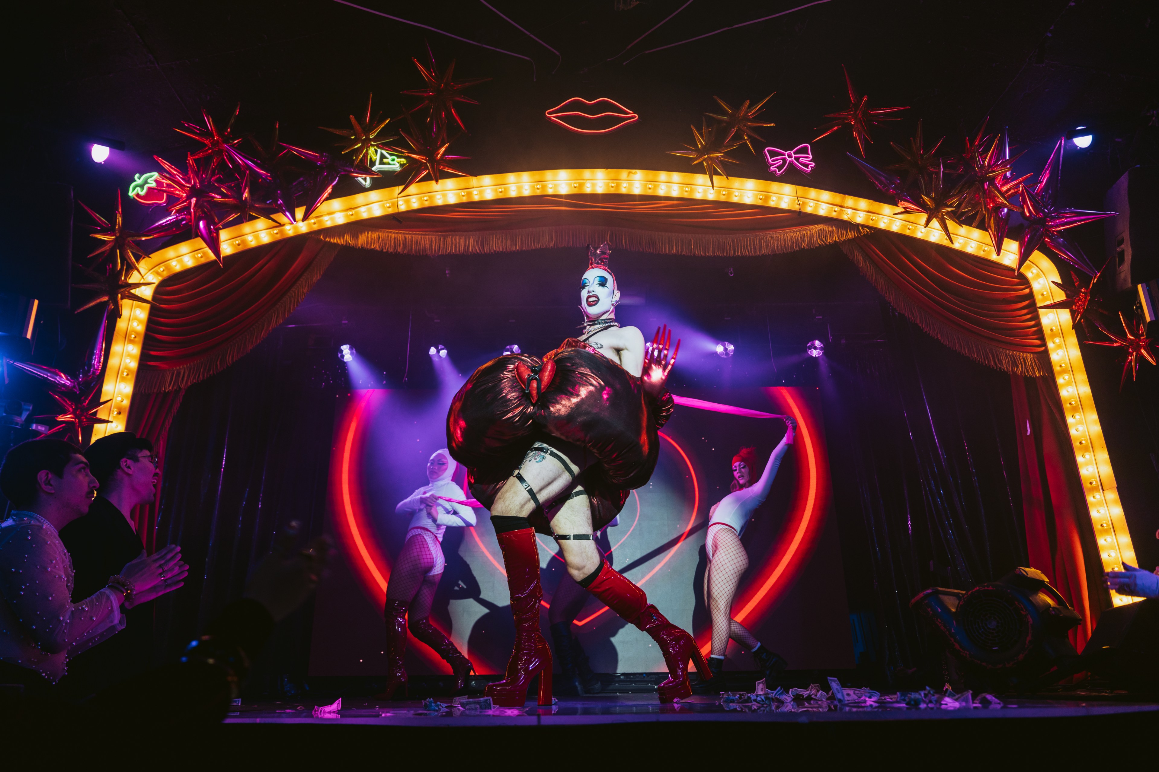 A performer in a theatrical setting, wearing a flamboyant outfit with tall red boots, is on stage in front of a vibrant, heart-themed neon backdrop.