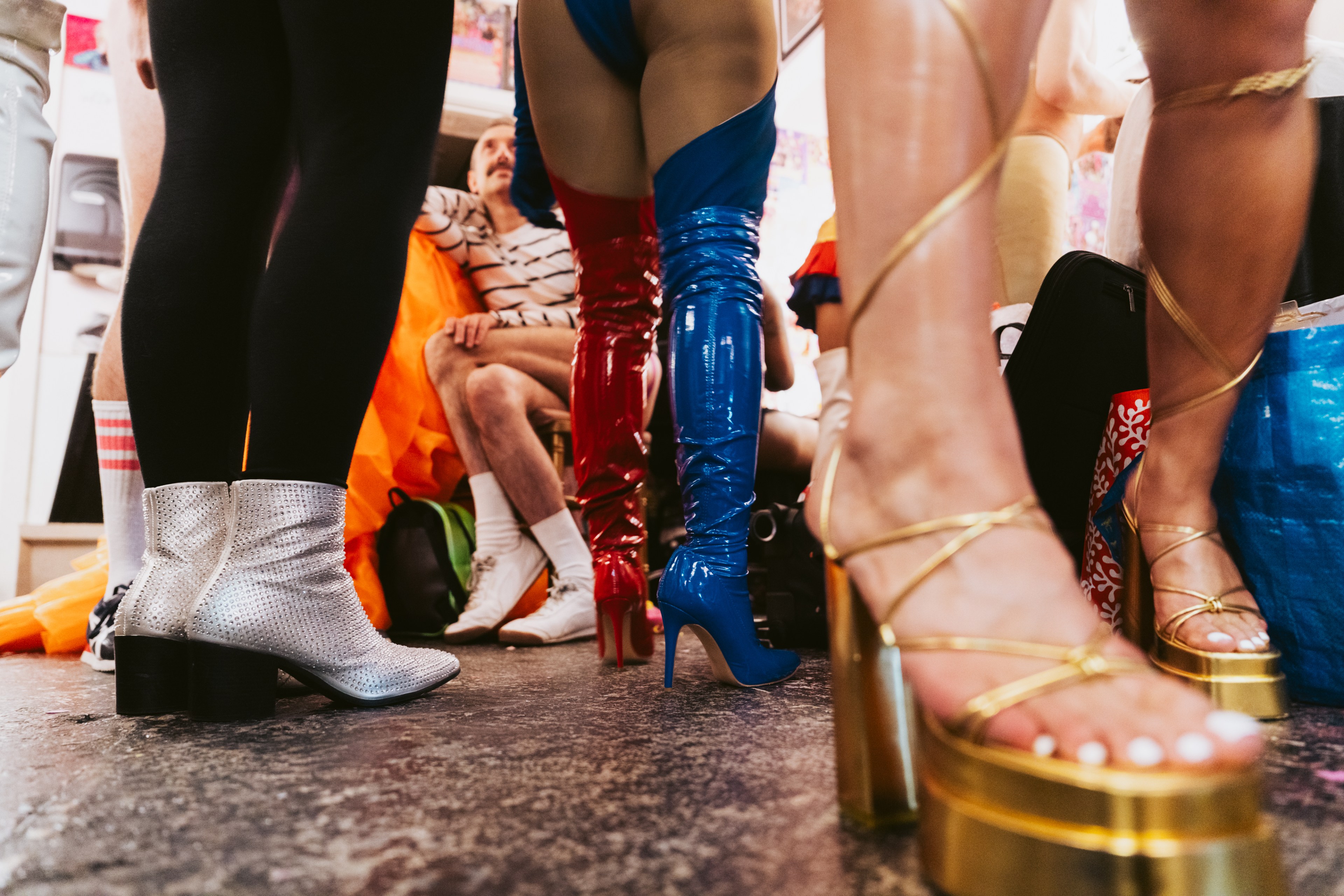 People wearing vibrant footwear gather indoors. Visible are shimmery silver boots, colorful thigh-high boots, and gold platform heels, with a seated person in the background.