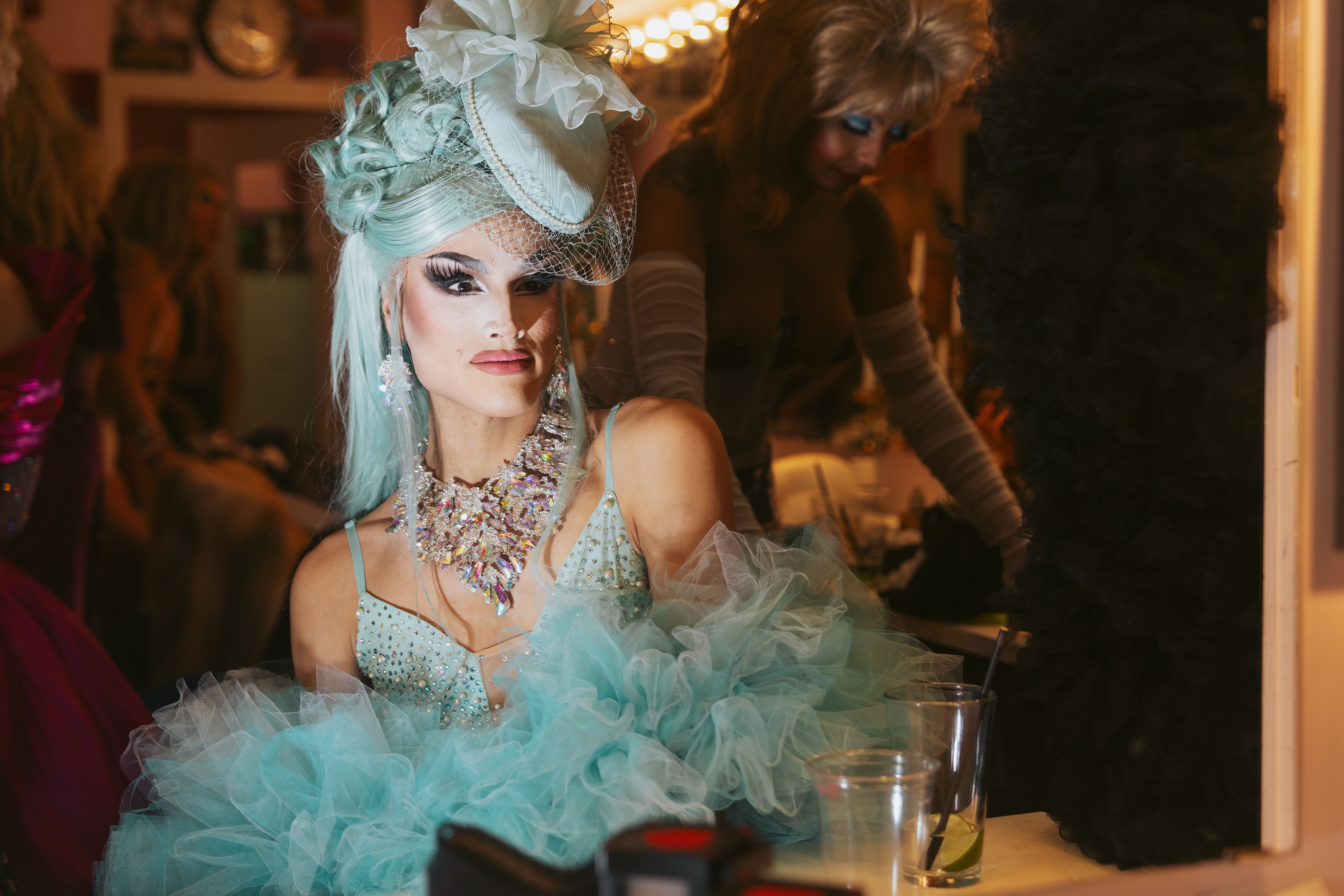 A person in dramatic makeup and a bright pastel outfit adorned with tulle and a flowery hat sits in a dressing room, with another person in the background.