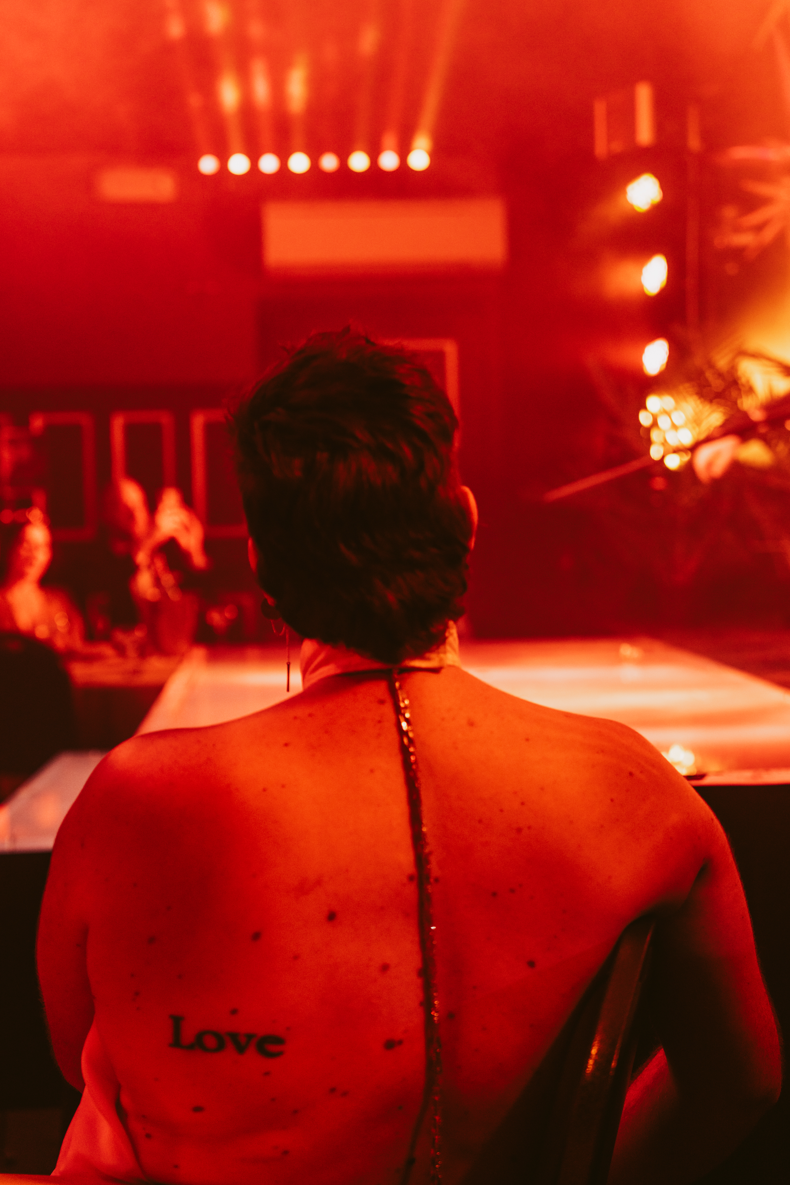 A person with short dark hair and a &quot;Love&quot; tattoo on their bare back is seated in a dimly lit room with red lighting and spotlights, likely at a performance event.