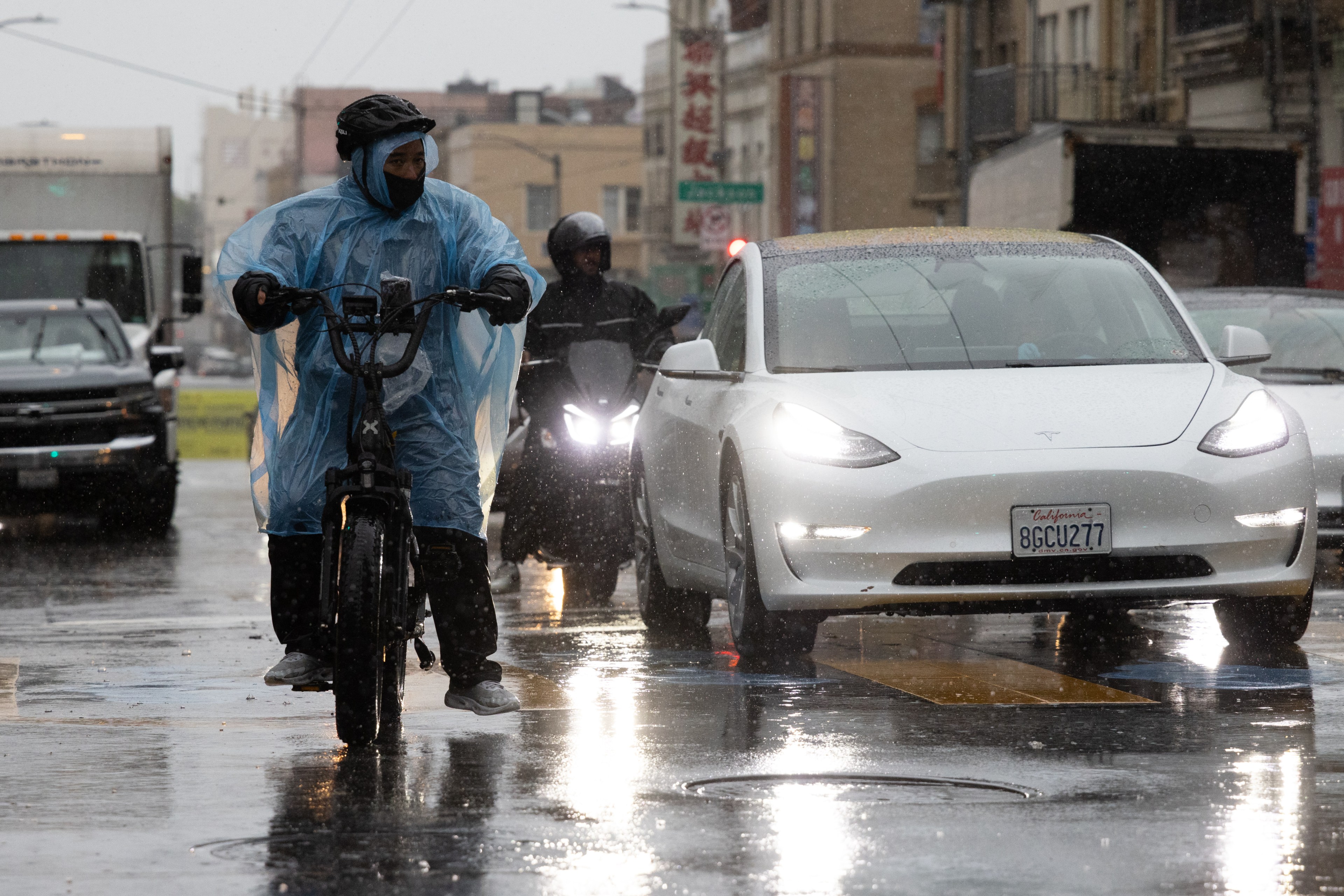 A person in a blue rain poncho rides a bicycle through a rainy street, while cars, including a white Tesla, drive beside him. Motorcyclists are also visible.