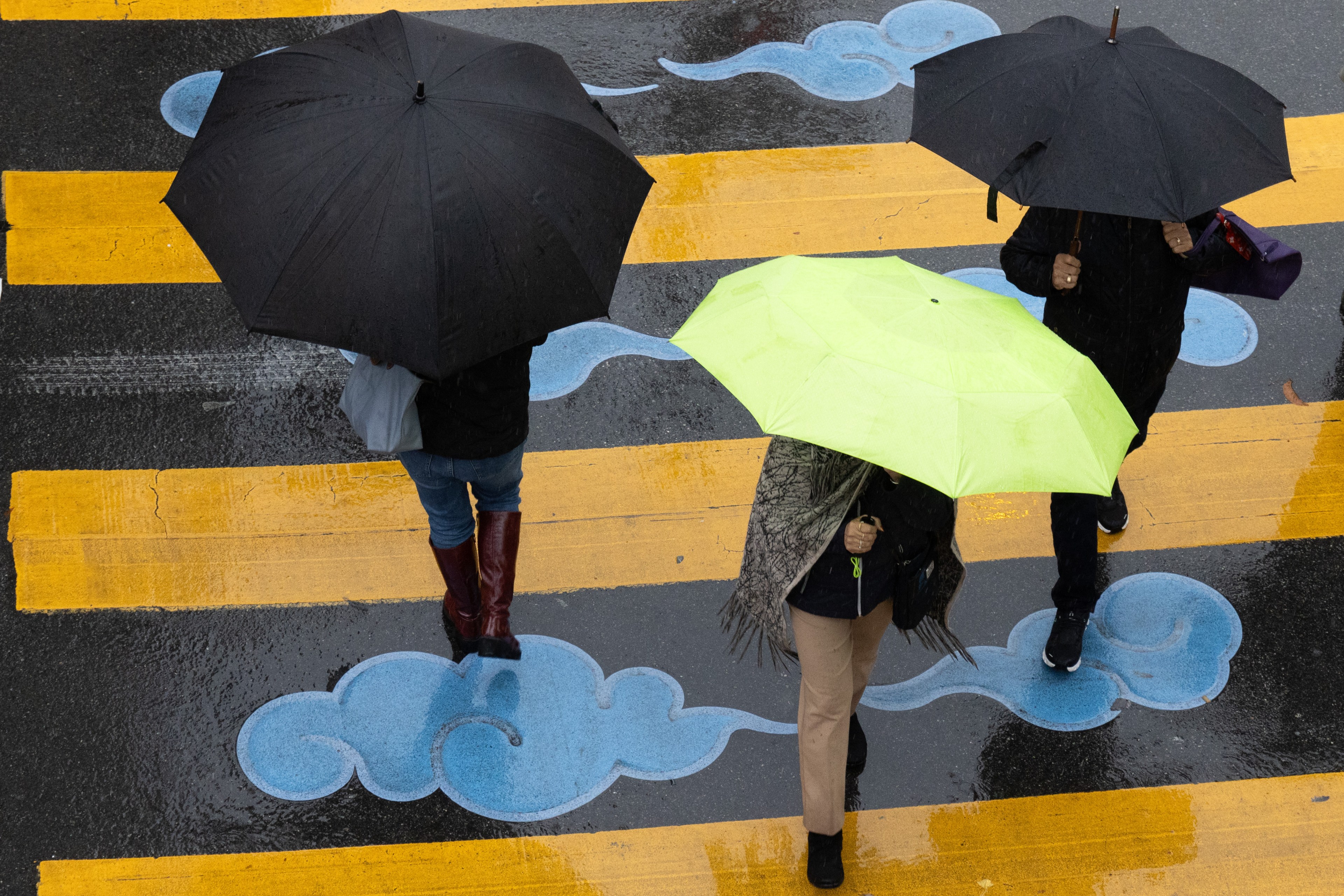 Three people with umbrellas—a lime green one and two black ones—cross a wet street painted with blue cloud designs and yellow stripes.