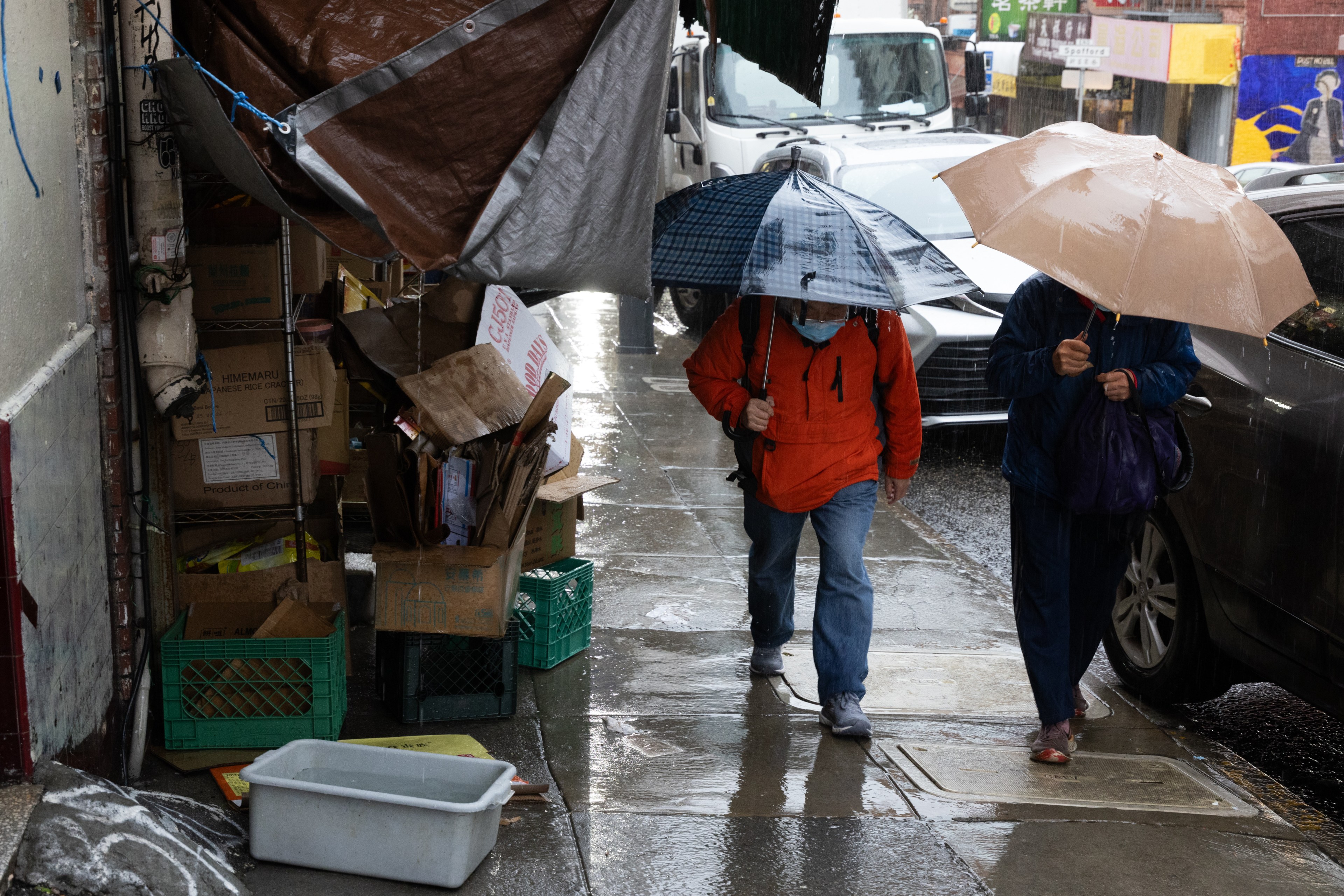 Two people are walking on a rainy street under umbrellas. Cardboard boxes and crates are stacked nearby, partially covered by a tarp.