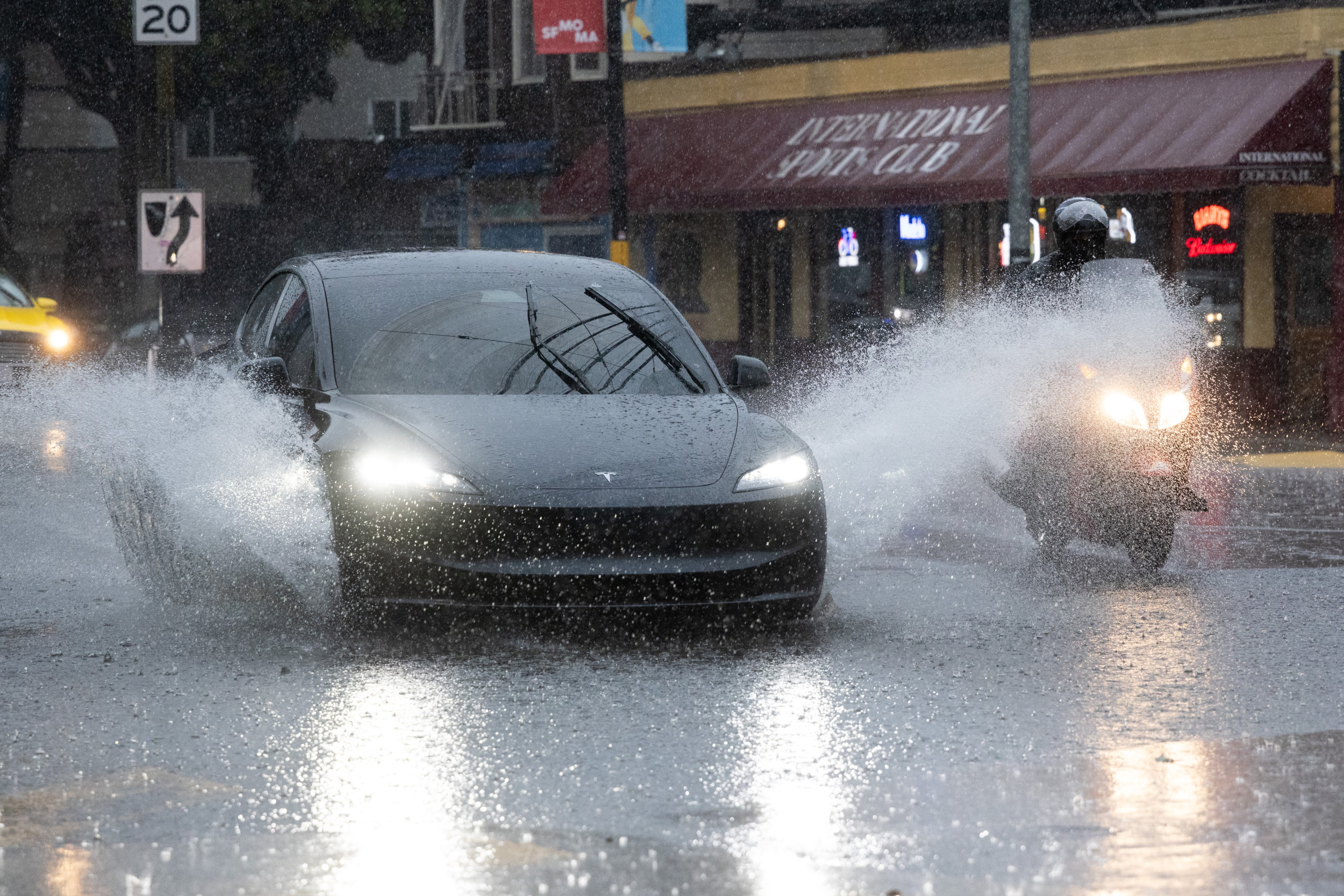 A black car and a scooter ride through a rain-drenched street, splashing water. A sign shows a 20 mph speed limit, and buildings with neon lights are in the background.