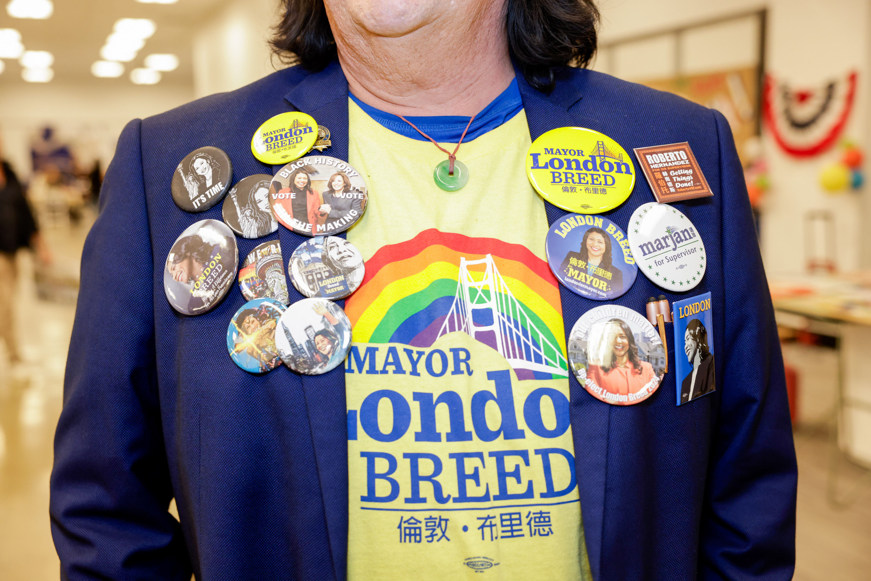 A person wears a blue jacket adorned with campaign buttons promoting Mayor London Breed, over a yellow shirt with a rainbow bridge graphic and text.