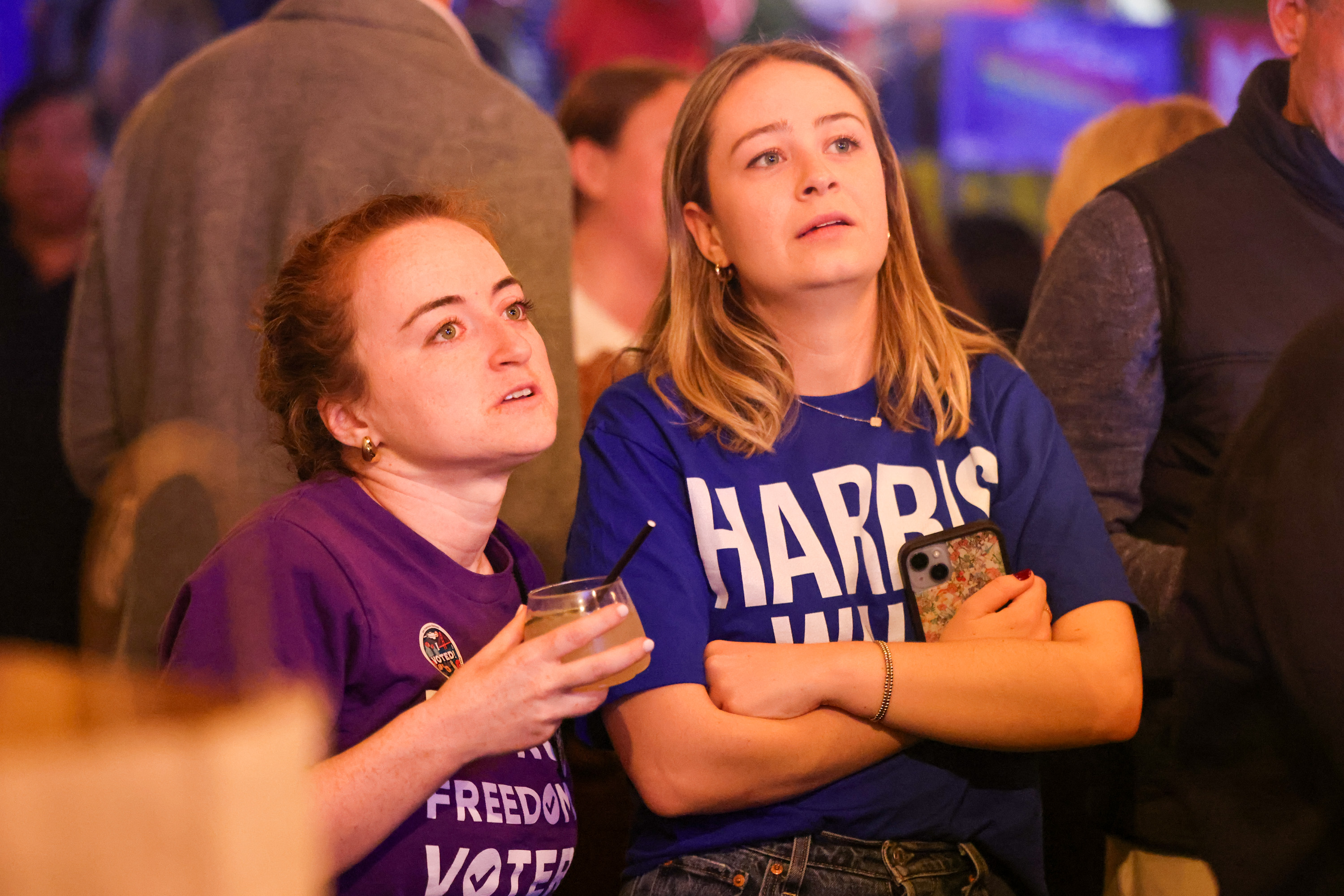 Two women stand in a crowded setting, looking intently at something off-camera. One holds a drink, and both wear shirts with text, including one that reads &quot;HARRIS.&quot;